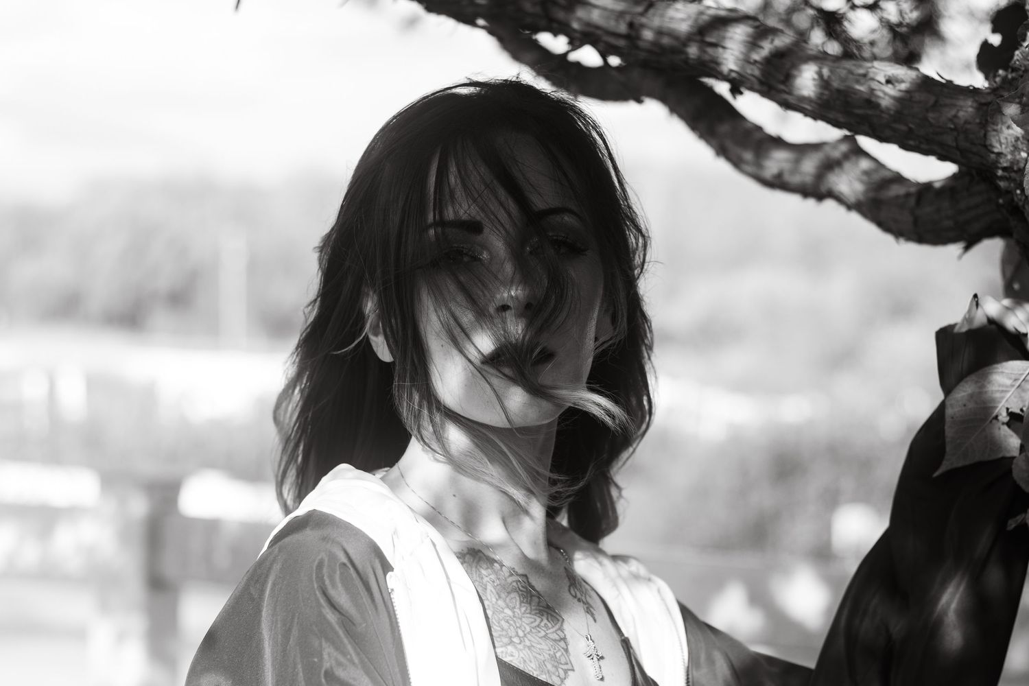 Black and white portrait of a dark-haired woman with wind-blown hair standing under a tree.