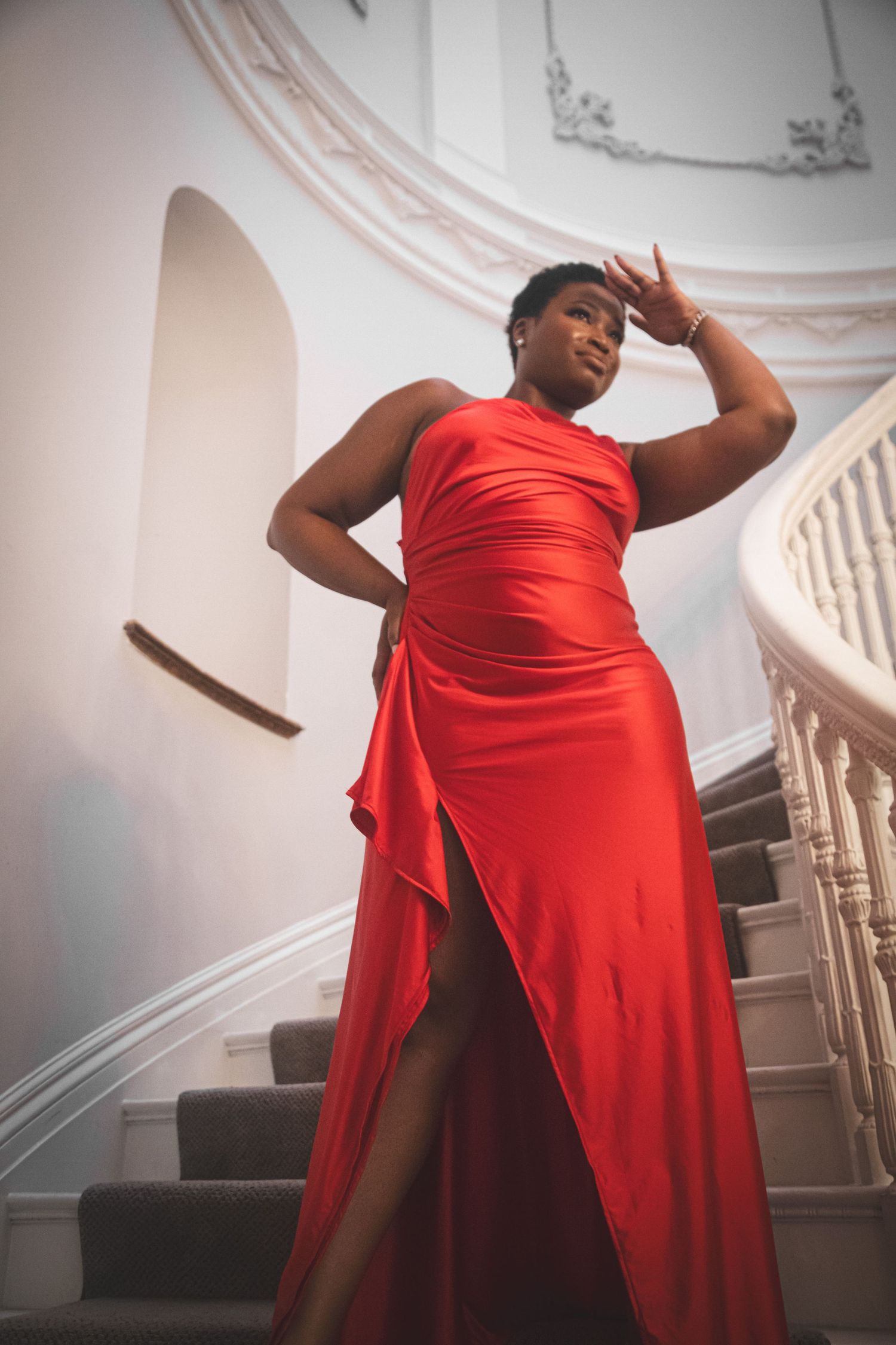 Black woman in a red one-shoulder satin gown with thigh slit poses on white ornate staircase.