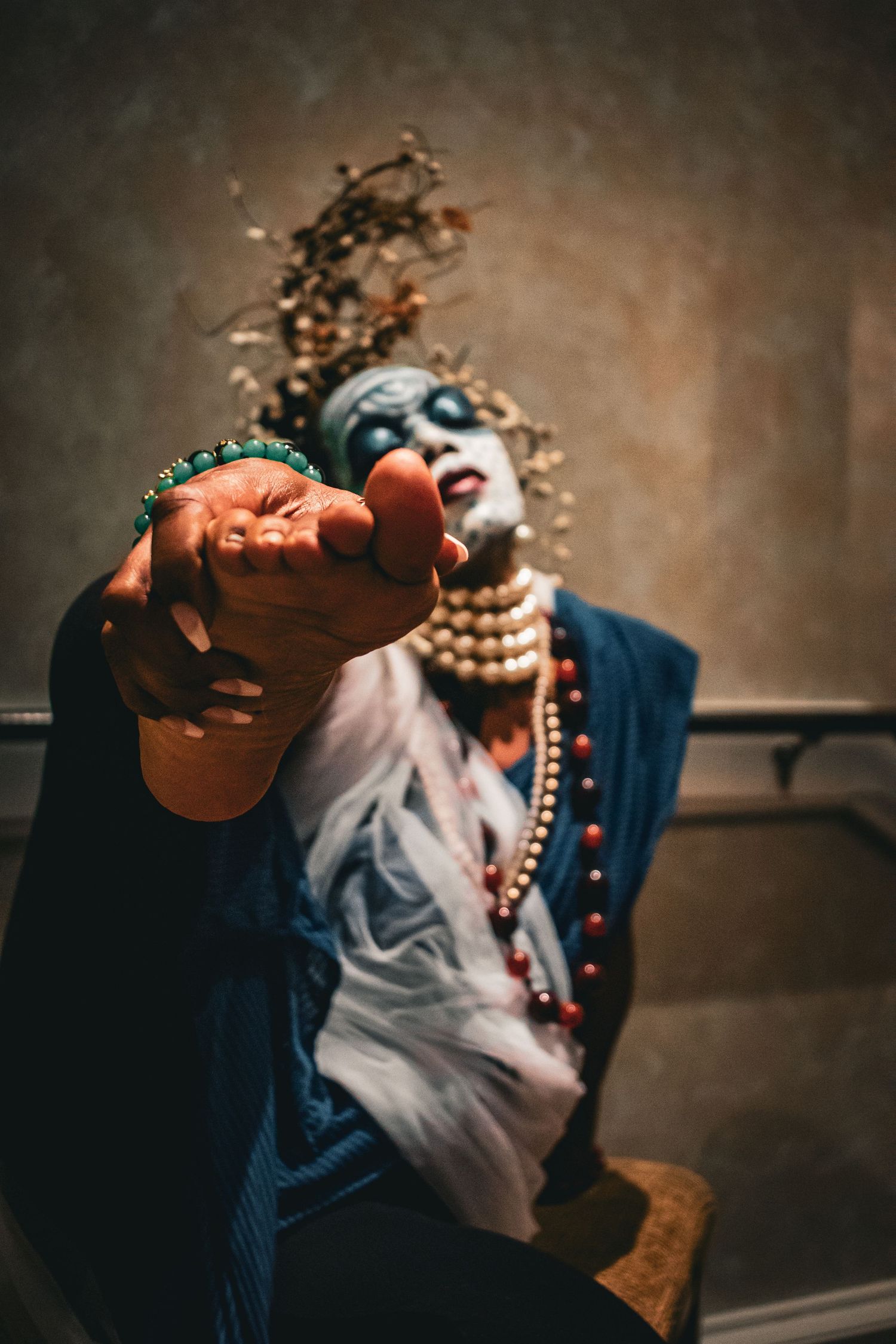 Performer in blue face paint and pearl necklaces extending bare foot toward camera dramatically.