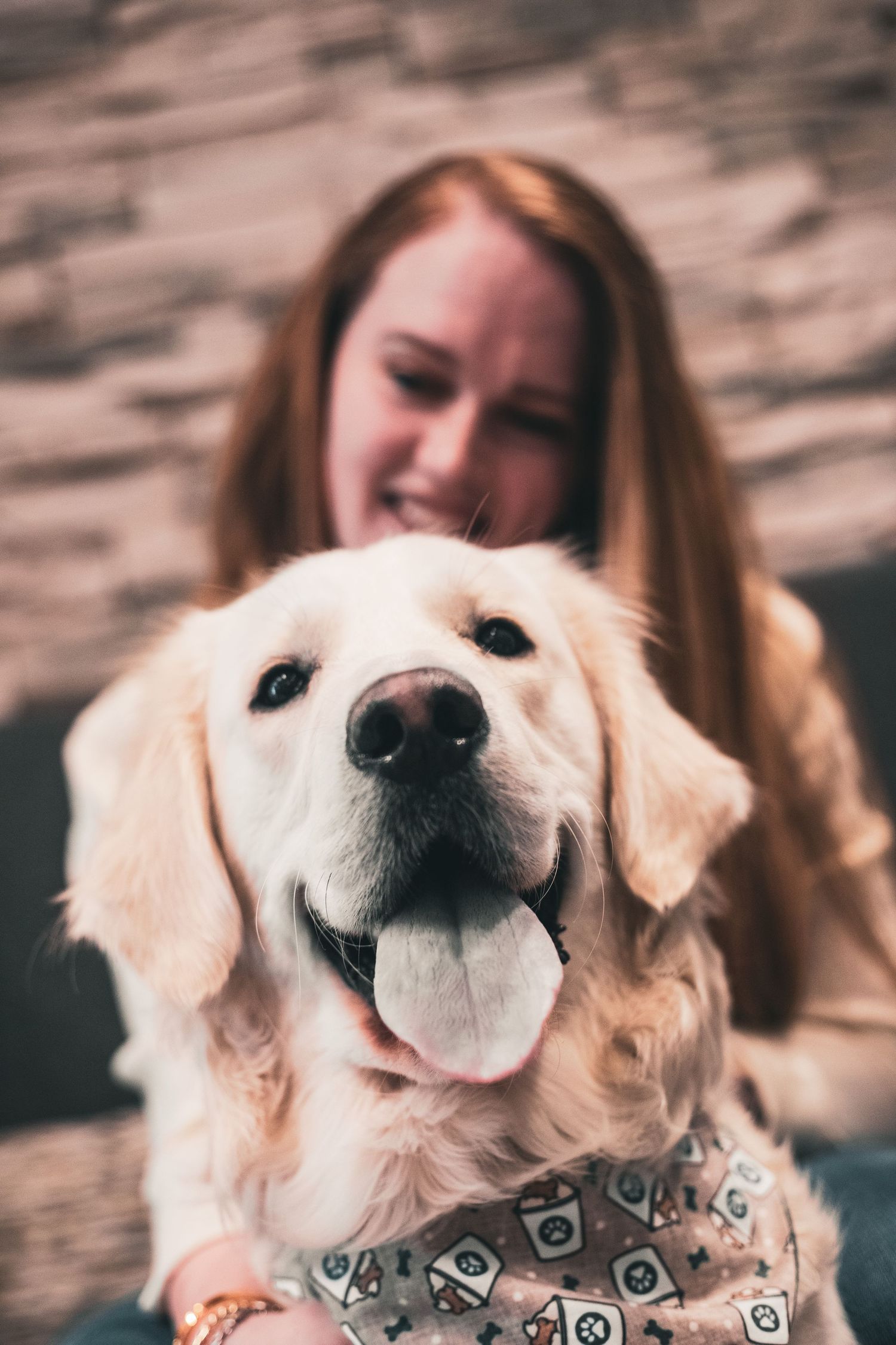 Smiling blonde woman petting a happy golden retriever wearing a bandana indoors.
