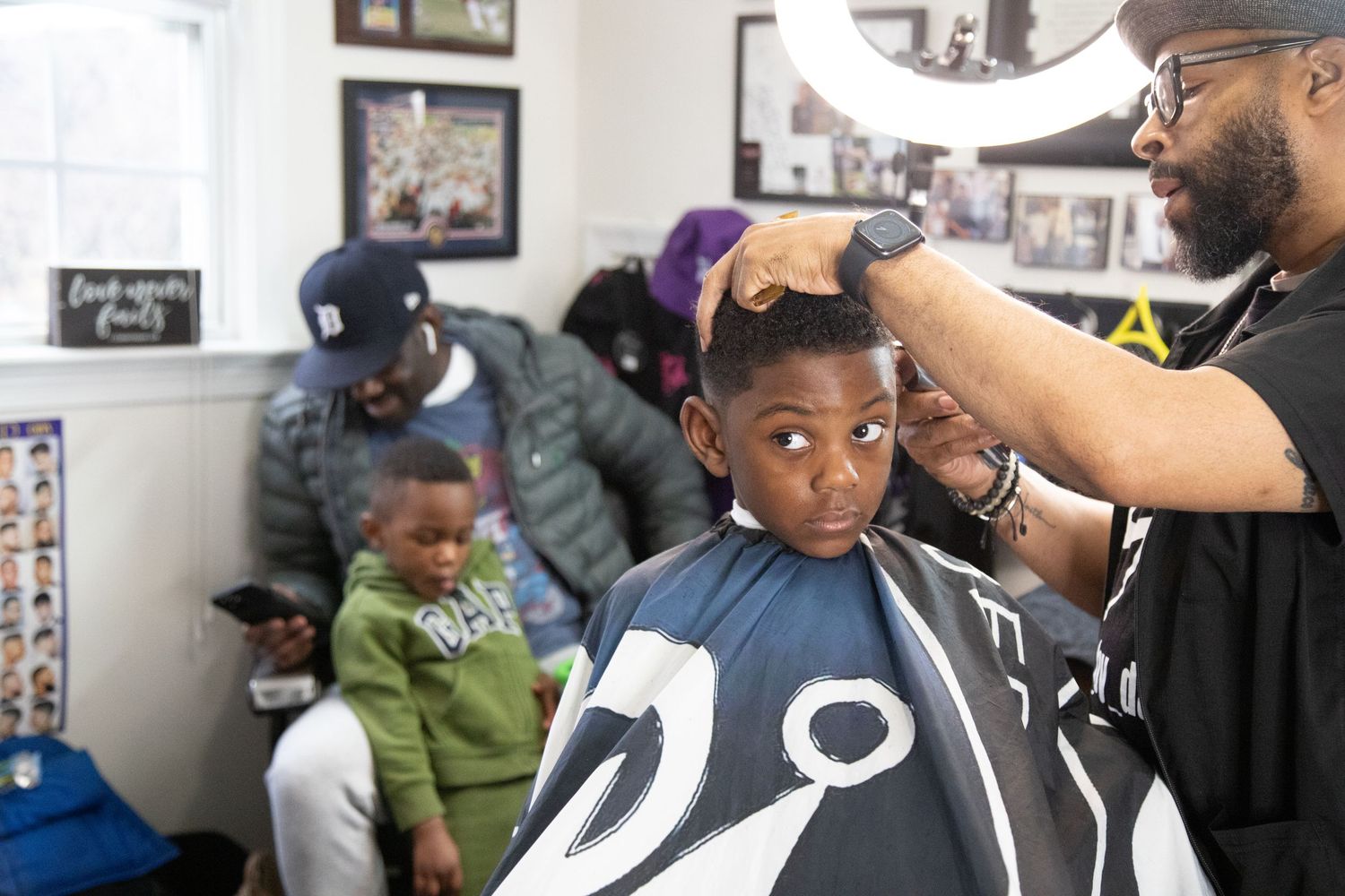 Young boy getting a haircut at a barbershop while family members wait in the background.