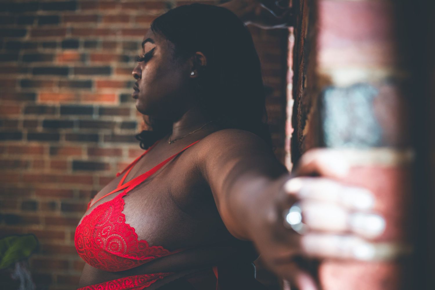 Woman in red lace bralette leaning against a brick wall in moody lighting.