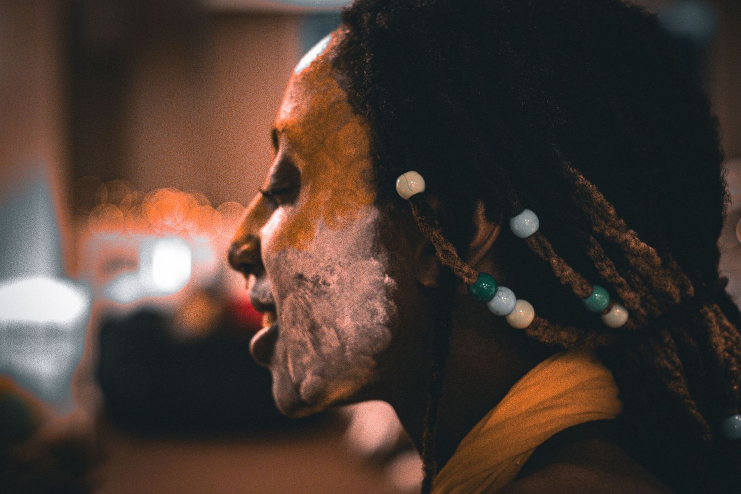 Person with dreadlocks adorned with colorful beads and white face paint in profile view.