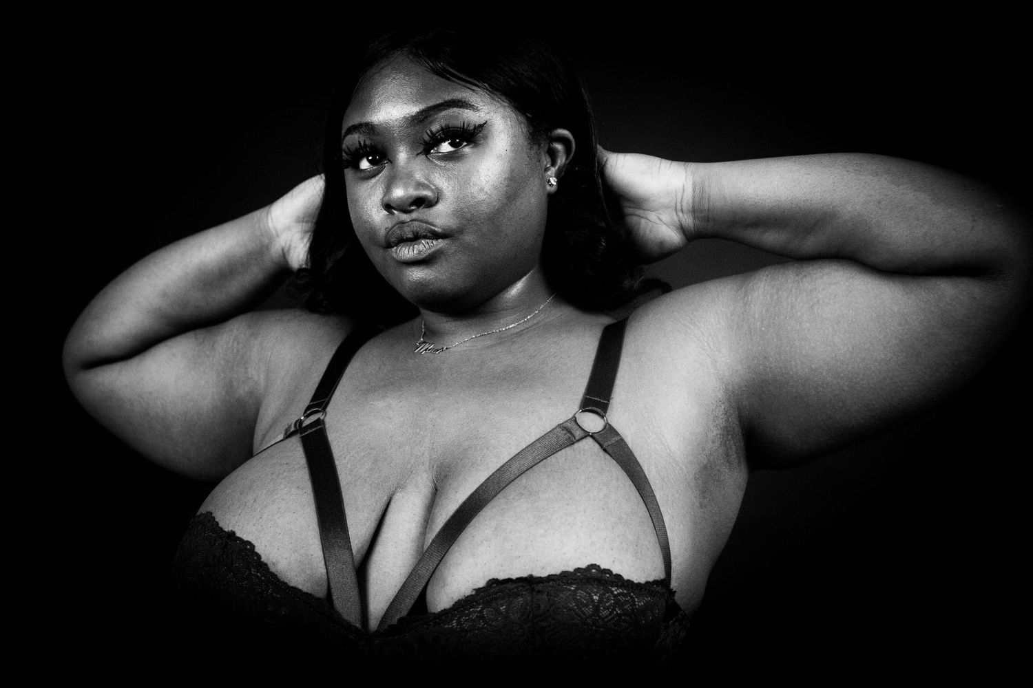 A Black plus-size woman poses in a black lace bralette with hands behind her head in a black and white photo.