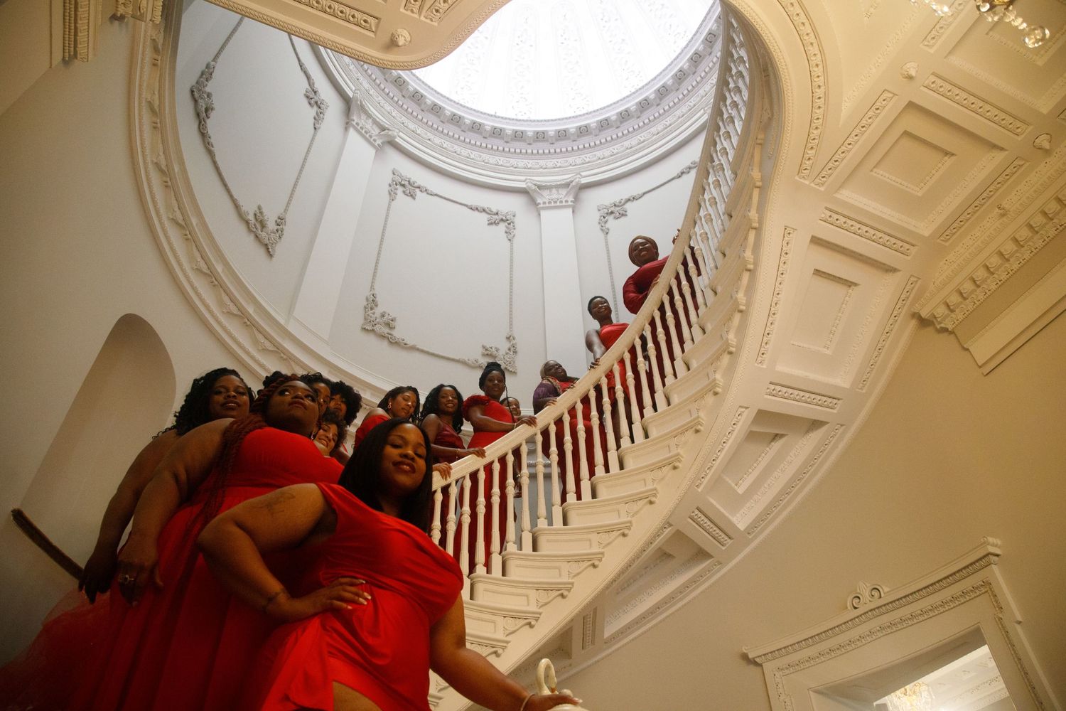Group of women in red dresses pose on a grand white spiral staircase in an elegant domed building.