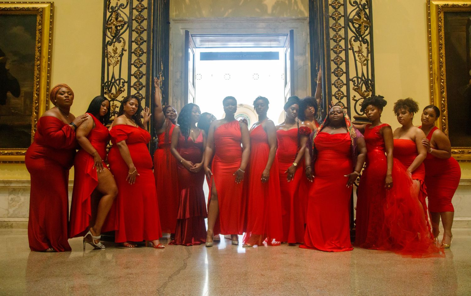 Large group of women in red formal gowns posing together in elegant marble hall.