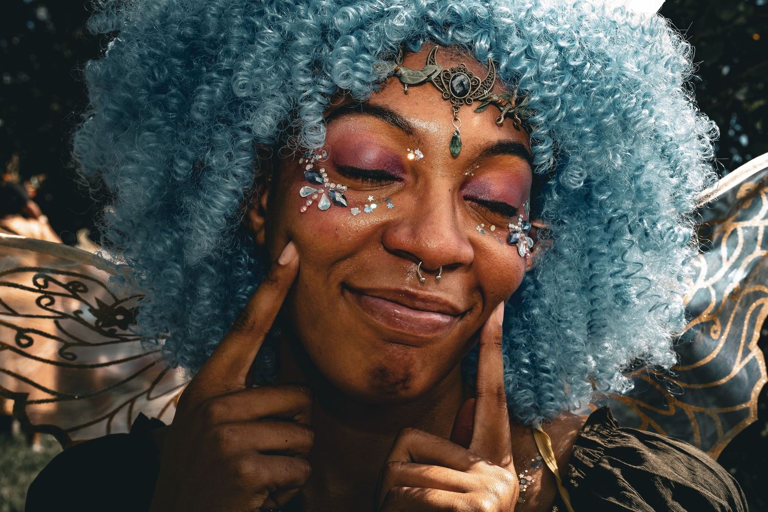 Close-up portrait of a woman with blue afro wig, glitter face gems, and fairy wings at a festival.