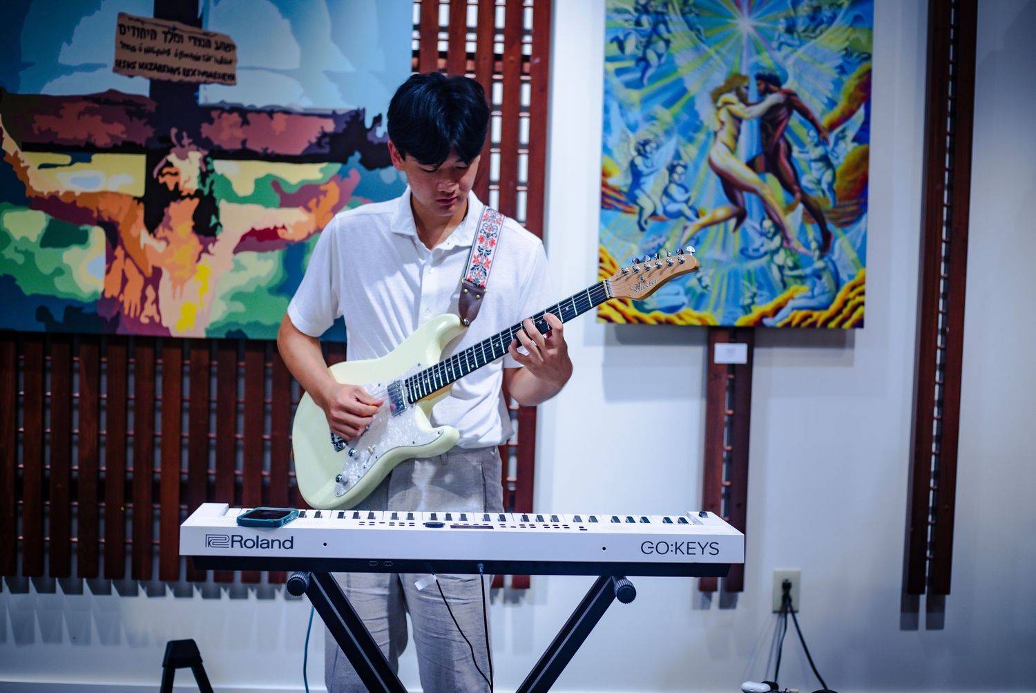 Young man playing white electric guitar at an art gallery with vibrant paintings displayed on the wall behind him.