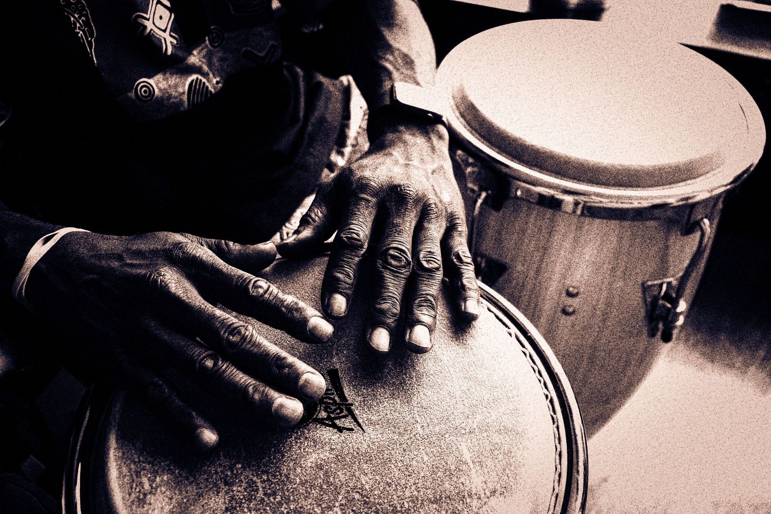 Close-up sepia-toned image of weathered hands playing traditional conga drums.