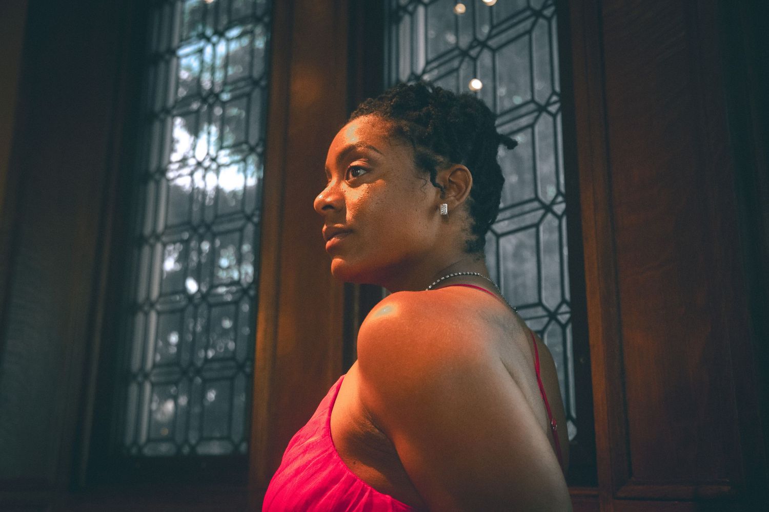 Black woman in a pink dress gazes thoughtfully sideways near a dark stained glass window in soft light.