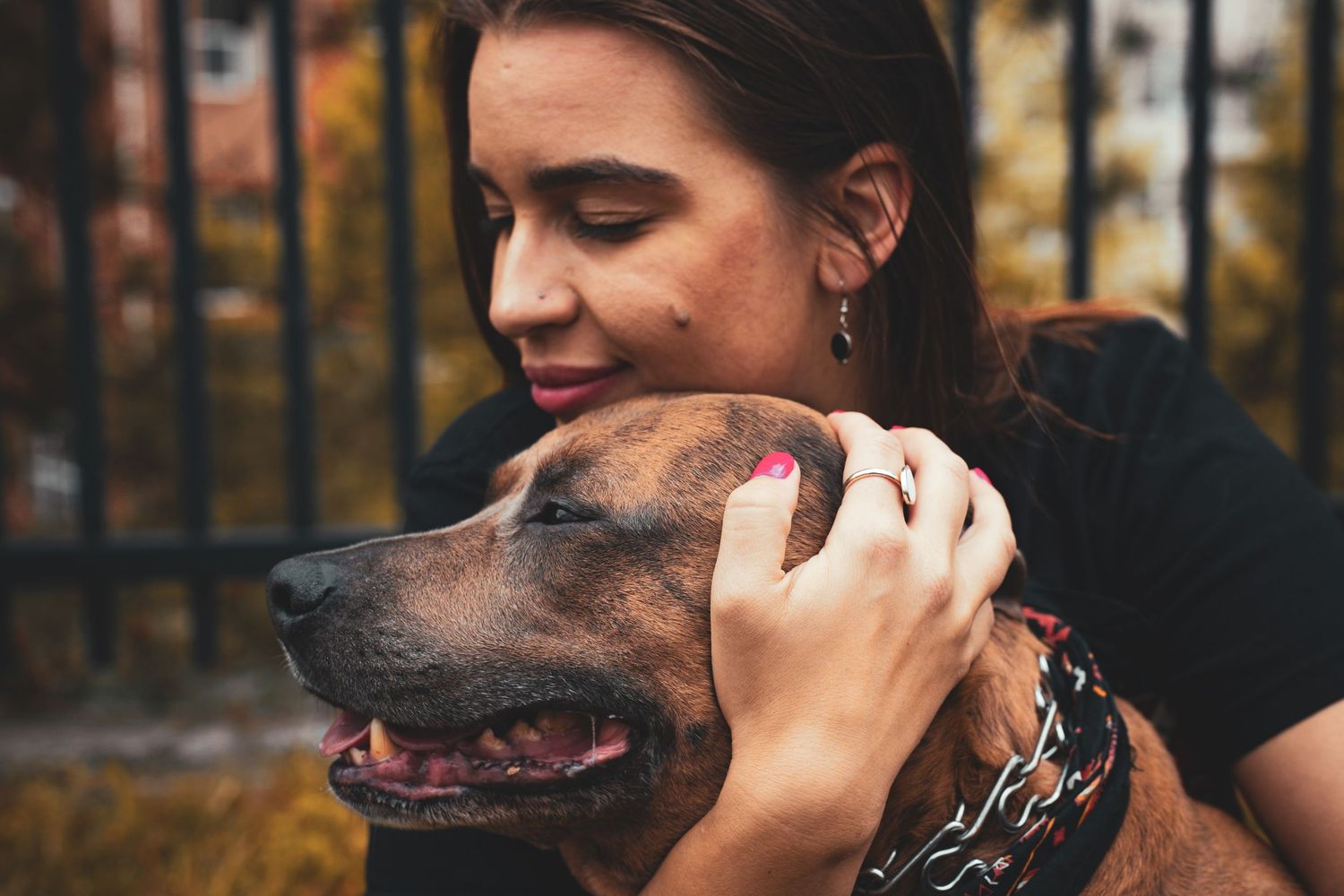 Woman with dark hair gently embracing a brindle, pitbull-mix dog near an outdoor black fence.
