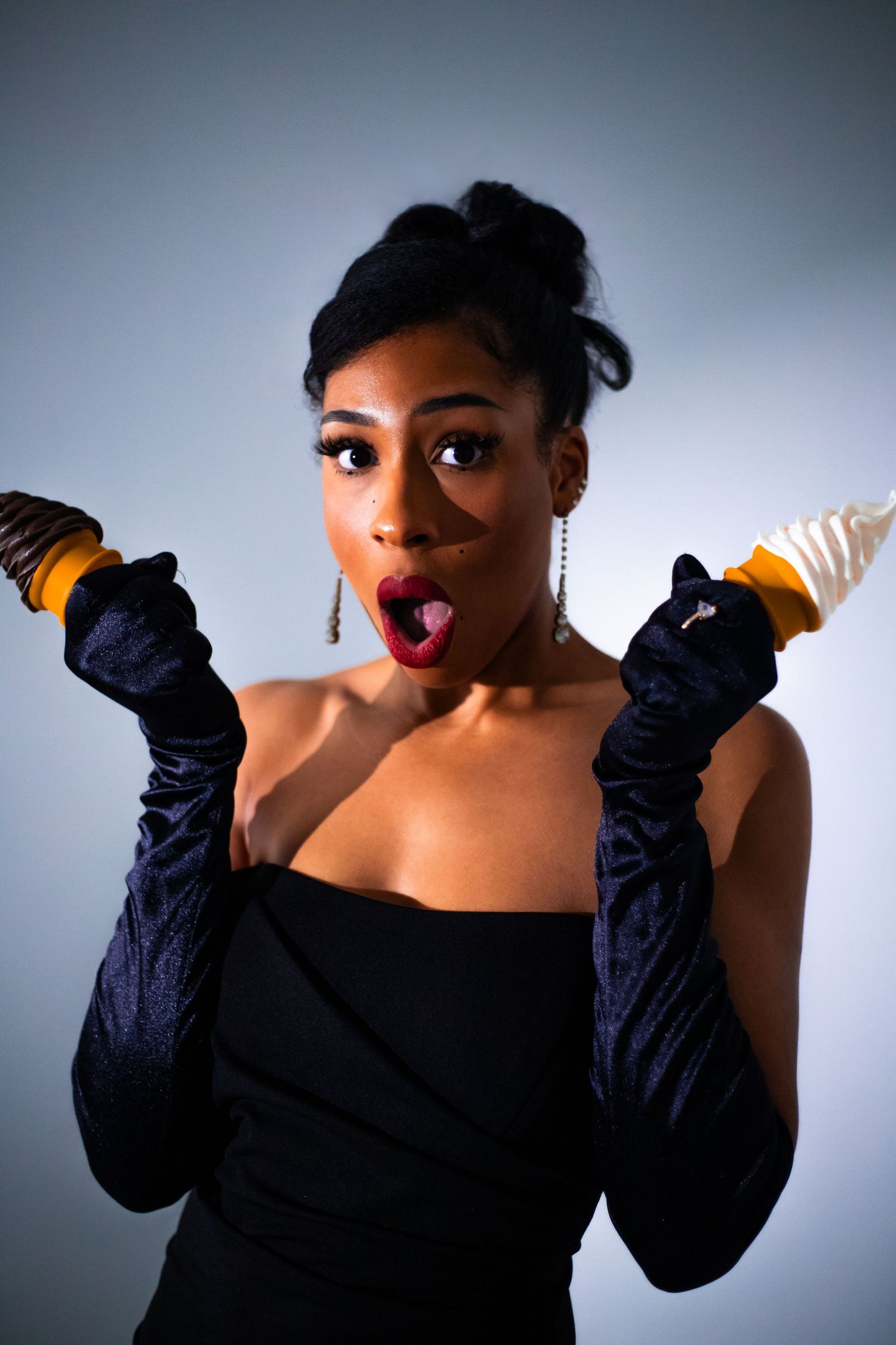 Glamorous woman in black dress and velvet gloves holding ice cream cones with a surprised expression.