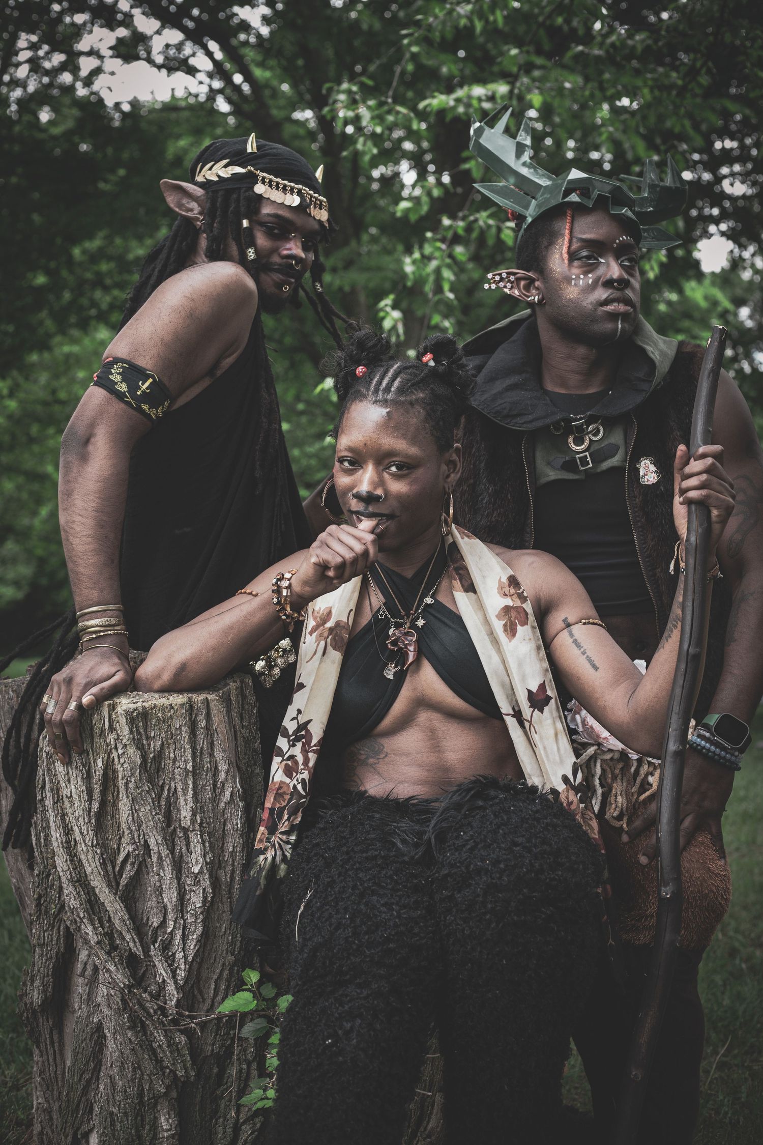 Three people in fantasy forest elf costumes with face paint and props pose among trees outdoors.