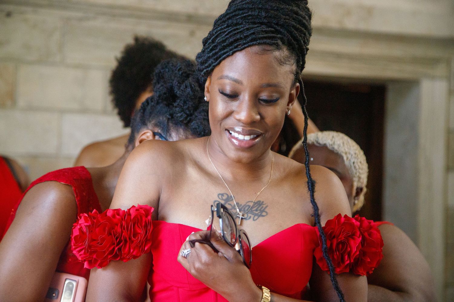 Smiling woman in red off-shoulder dress with floral details looks at her phone indoors at an elegant event.