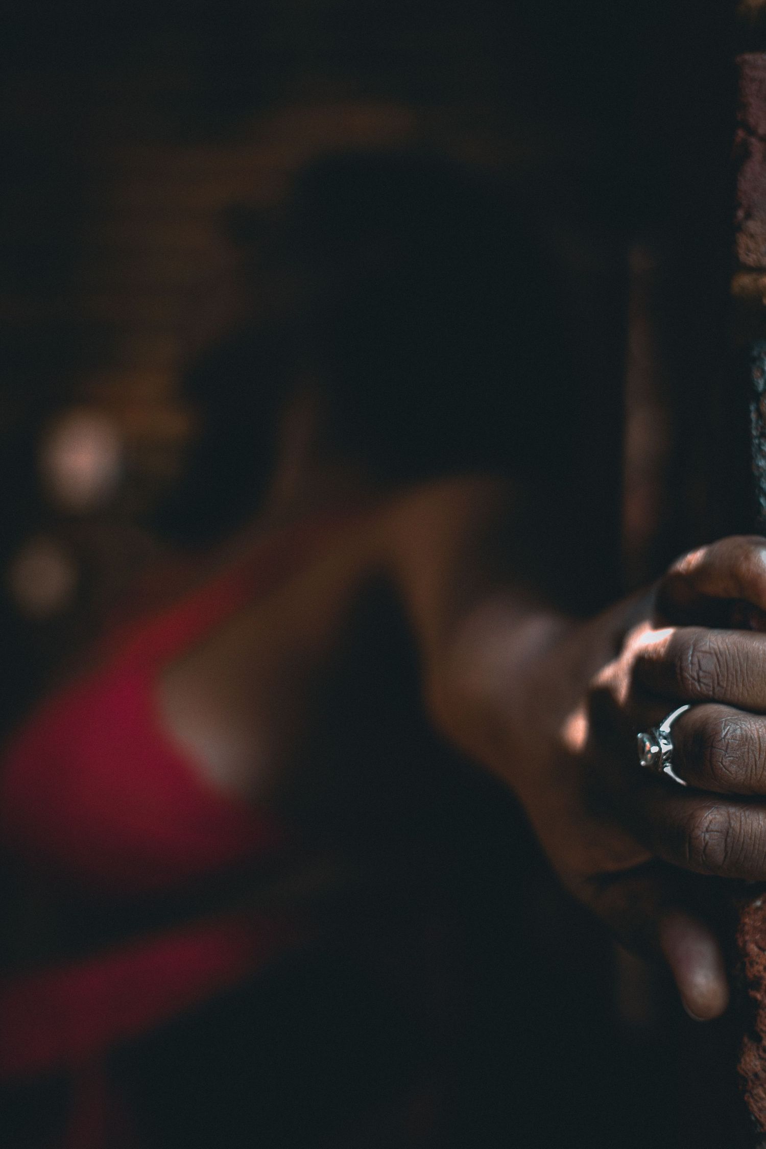 A close-up of a hand gripping a dark surface with a diamond ring, blurred red lingerie figure in background.