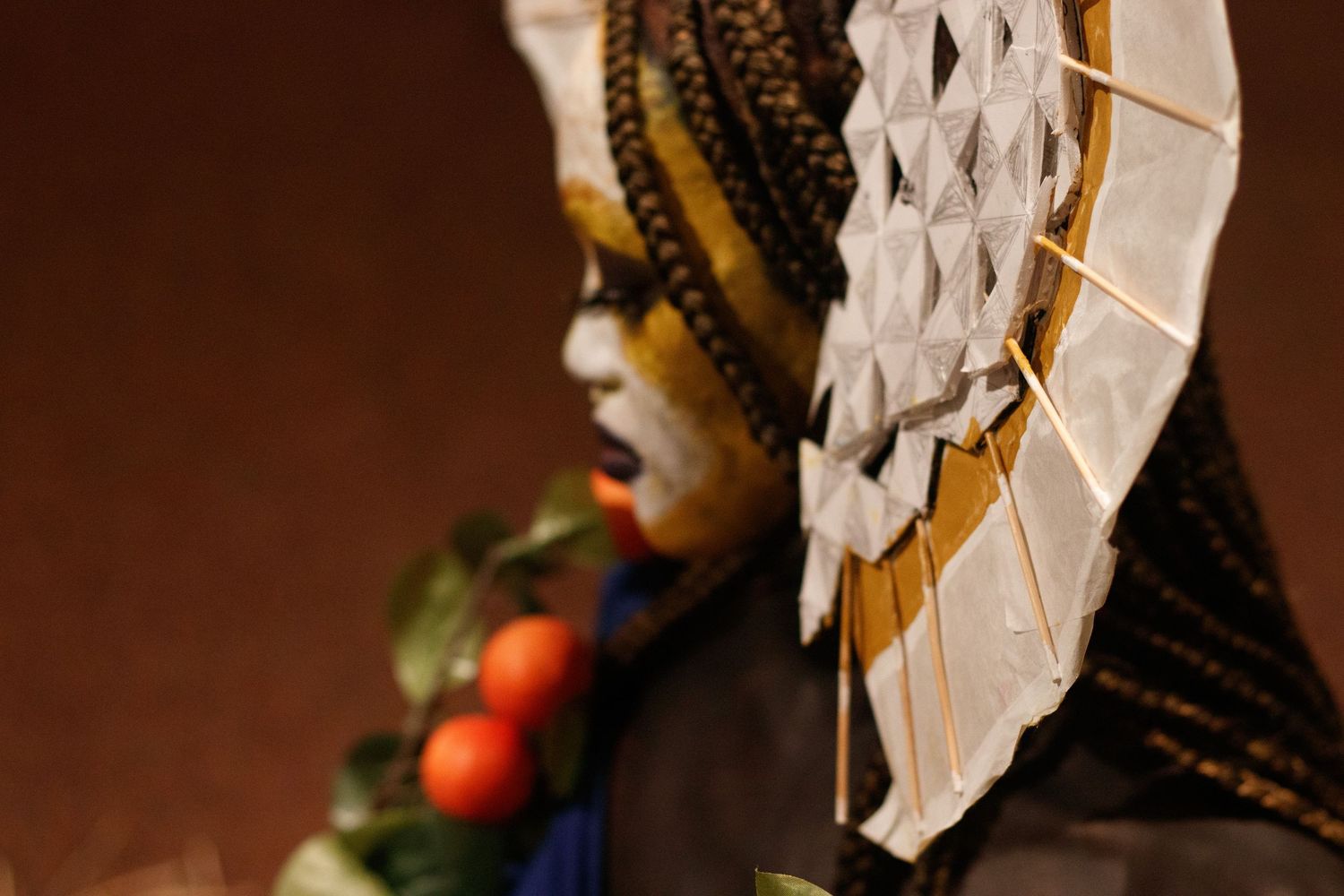 Performer in white face paint with decorative paper fan headdress and fruit accessories in profile.