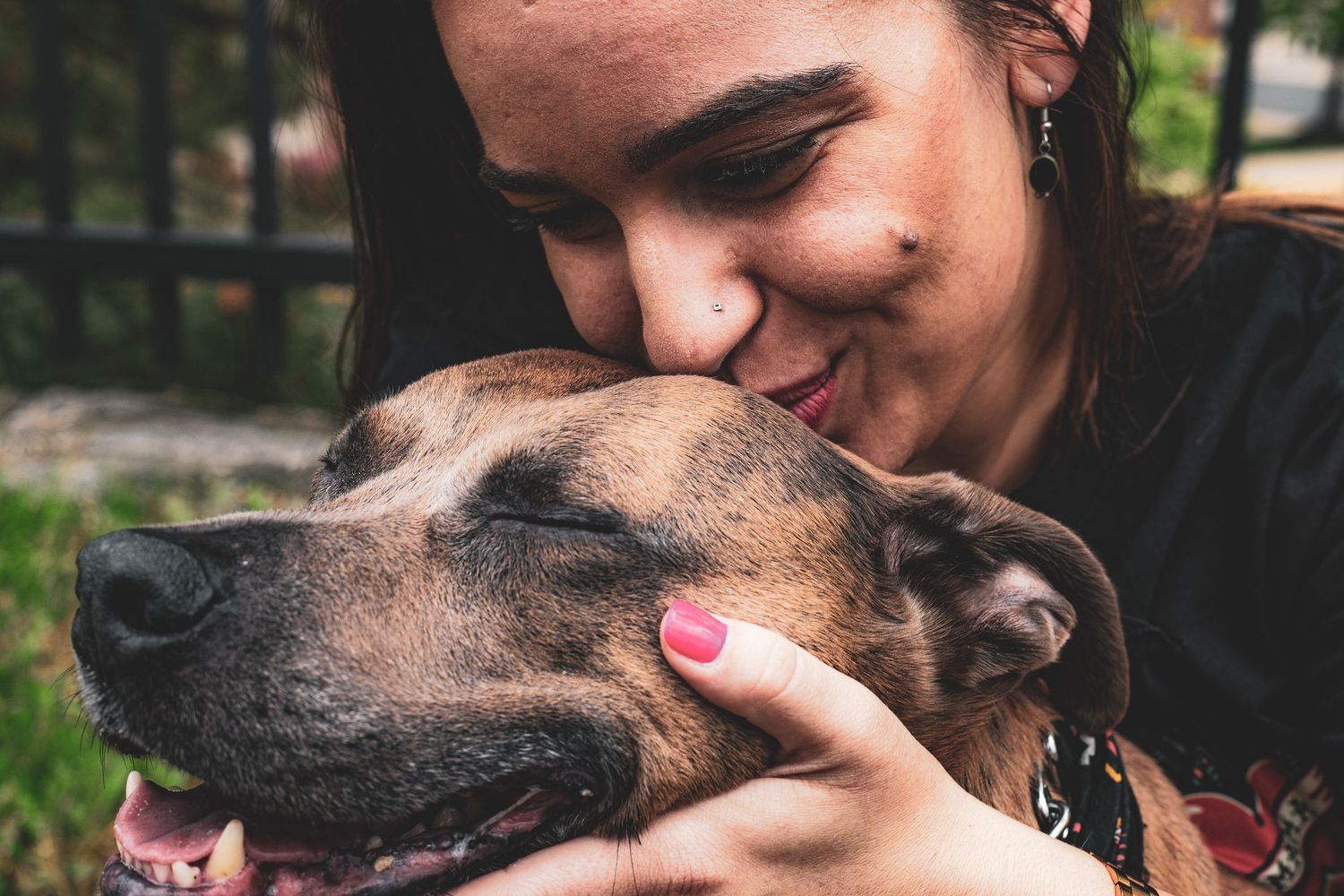 Close-up of a woman smiling and cuddling a brindle, pitbull-mix dog with pink nails visible.
