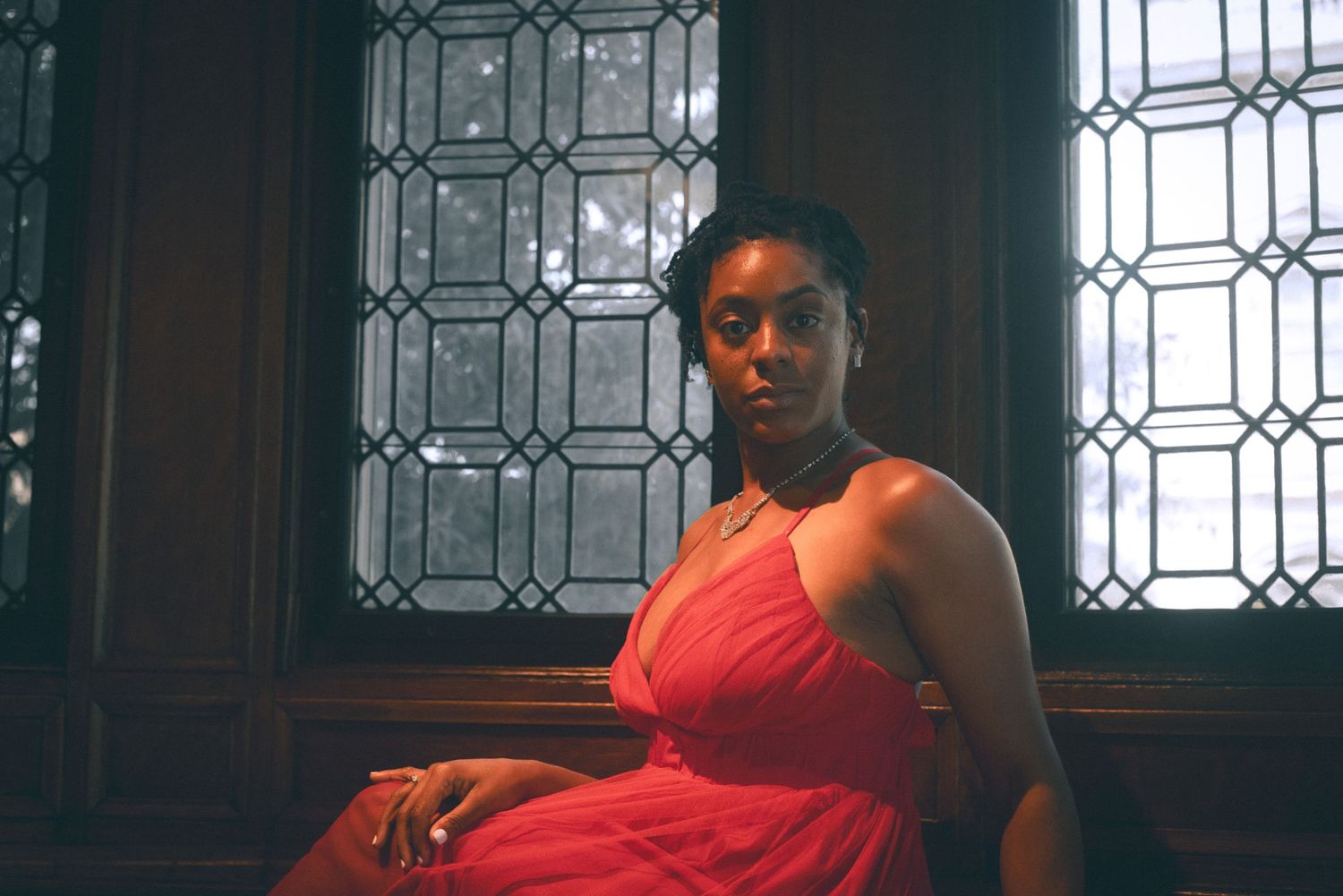 Young Black woman in a coral halter dress sits pensively near a decorative stained glass window.