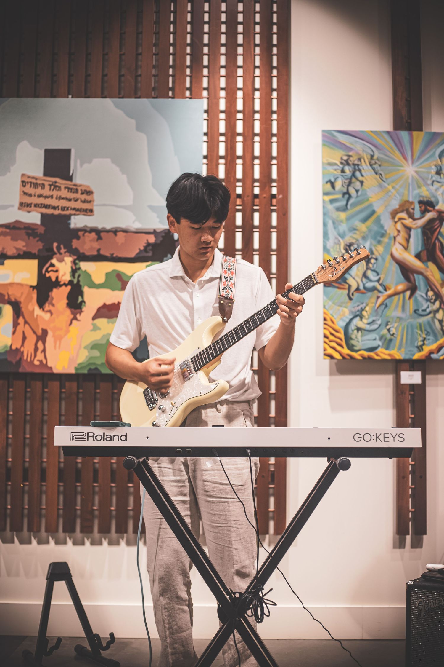 Young Asian man playing electric guitar in front of colorful paintings with a Roland GO:KEYS keyboard nearby.