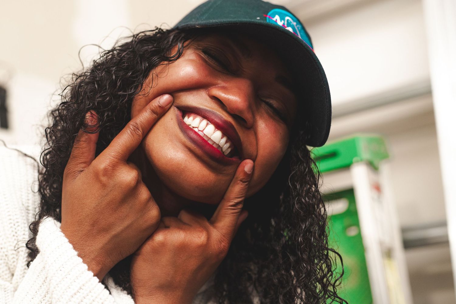 Joyful Black woman with curly hair and teal cap smiling broadly, touching her cheeks with one hand.