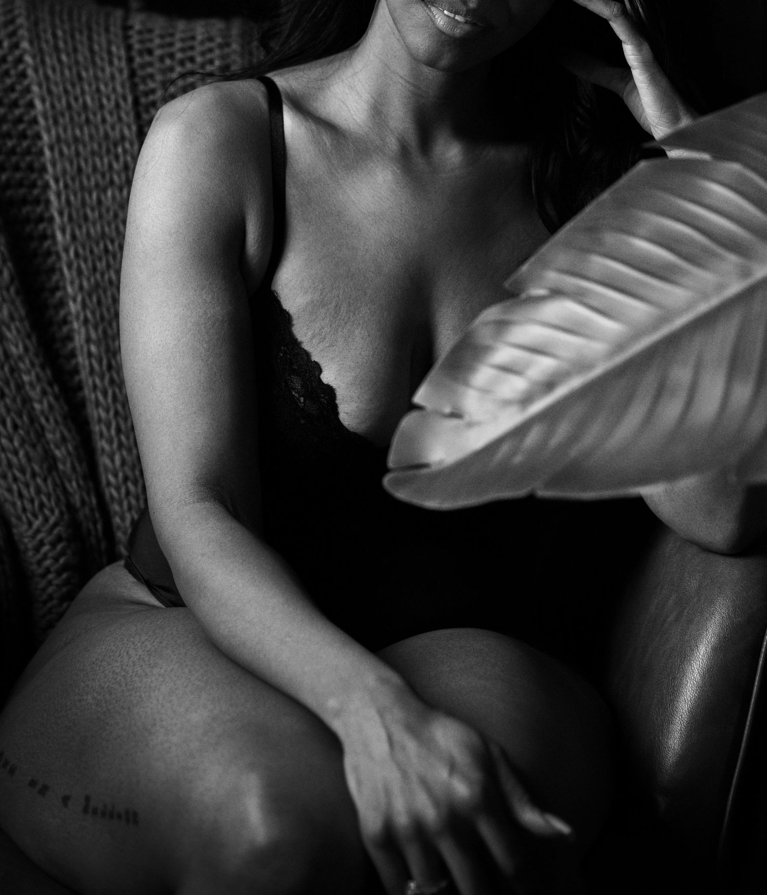 Black and white boudoir photo of a woman in black lingerie seated on a leather chair holding a leaf.
