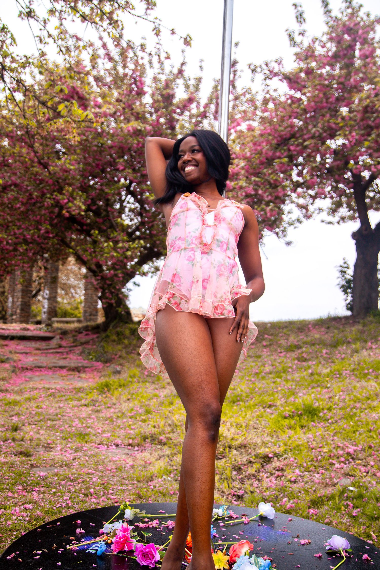 Smiling woman in pink floral outfit posing outdoors among blooming pink cherry blossom trees.