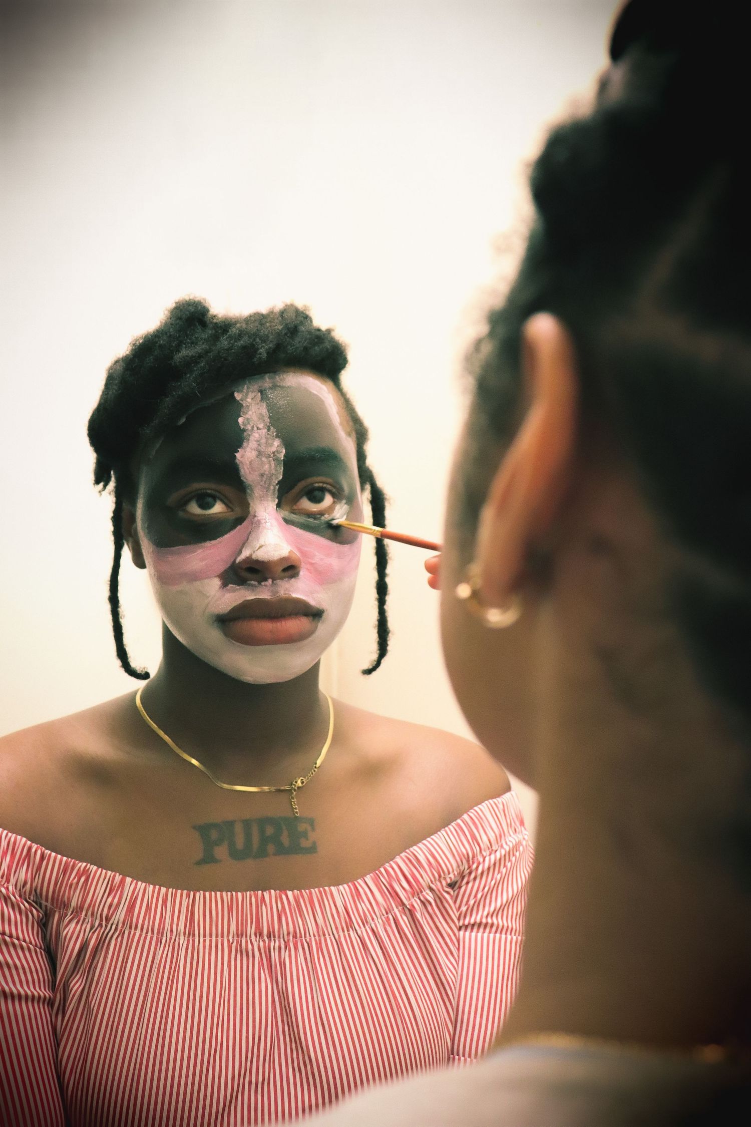 Artist applying colorful face paint to a young Black woman with braids wearing a red striped top and gold PURE necklace.