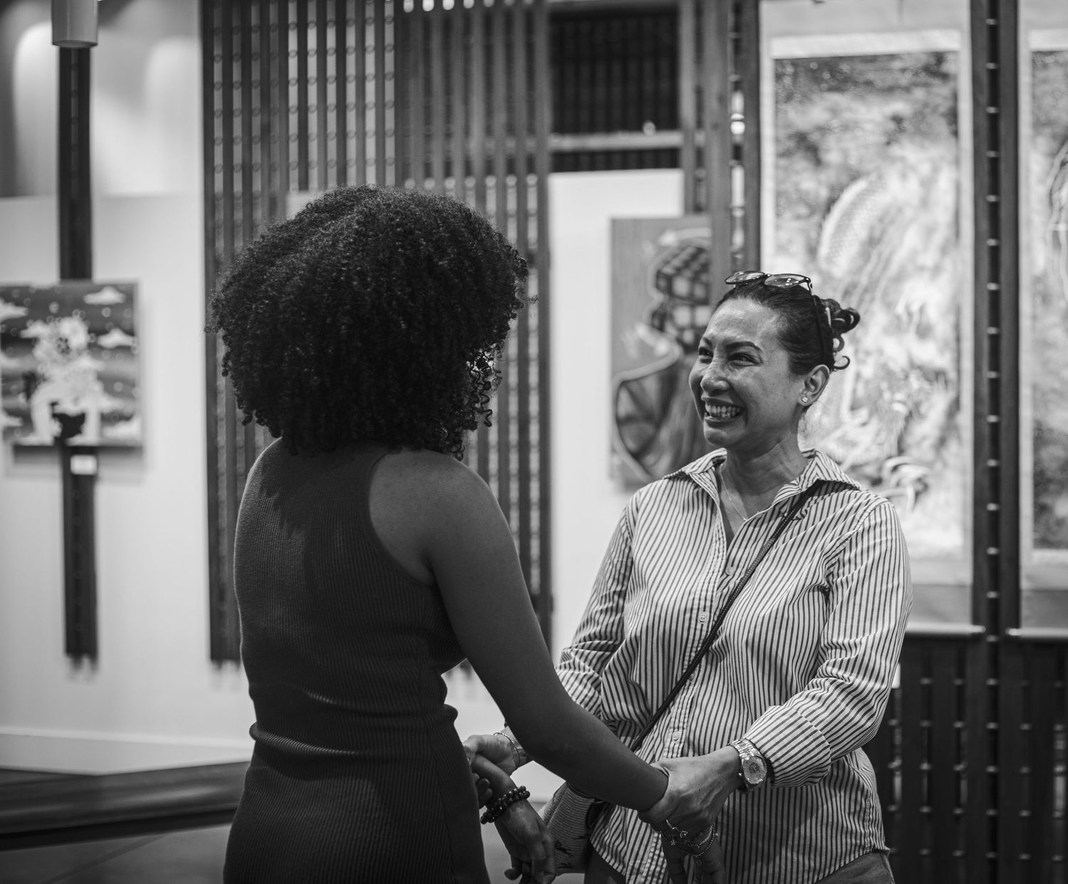 Two women smiling and shaking hands at an art gallery event in black and white photography.