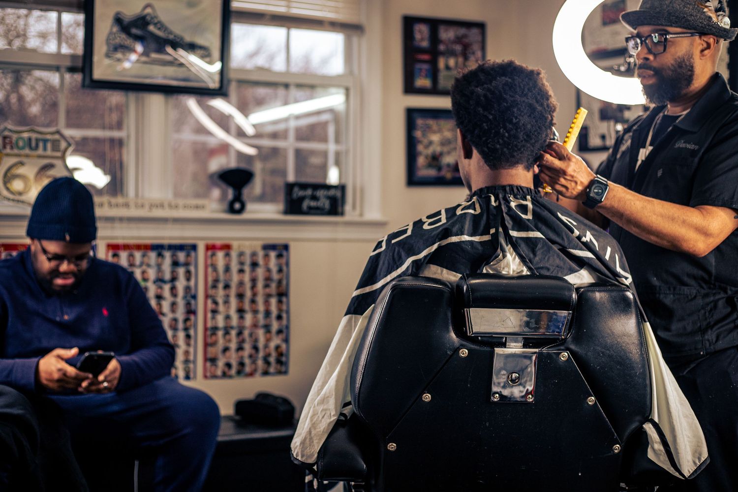 Barber styling a clients hair in a cozy barbershop while another customer waits on his phone.