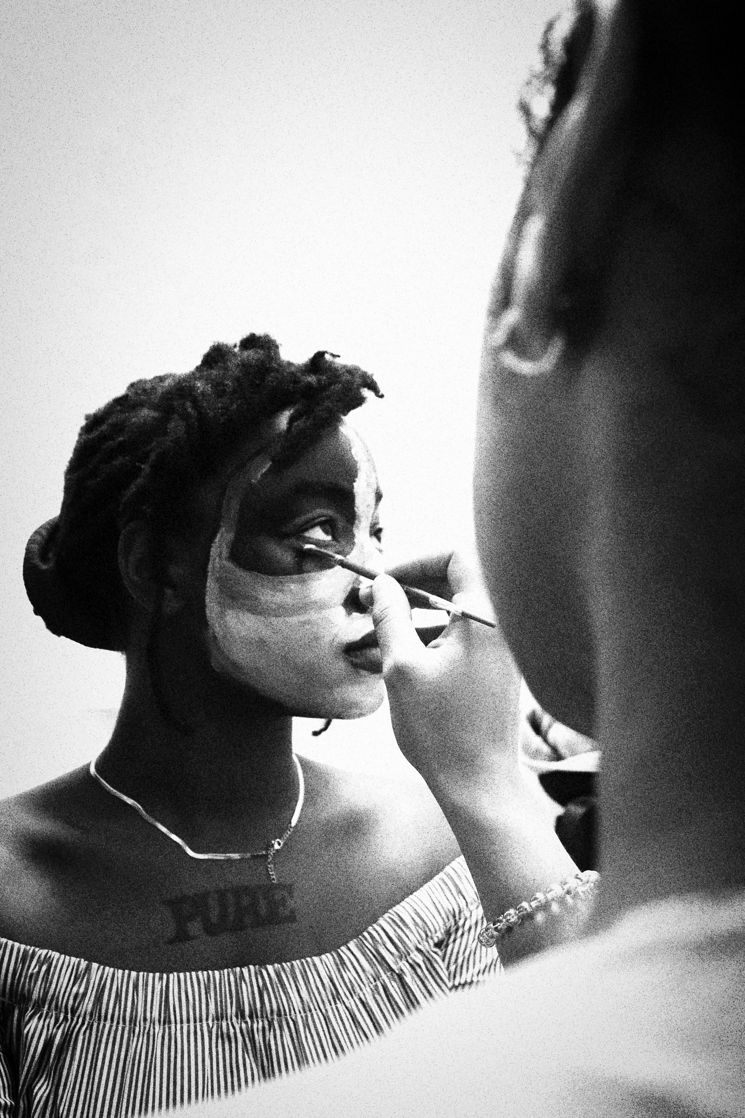 Black and white photo of a woman having face paint applied by an artist in a mirror reflection.