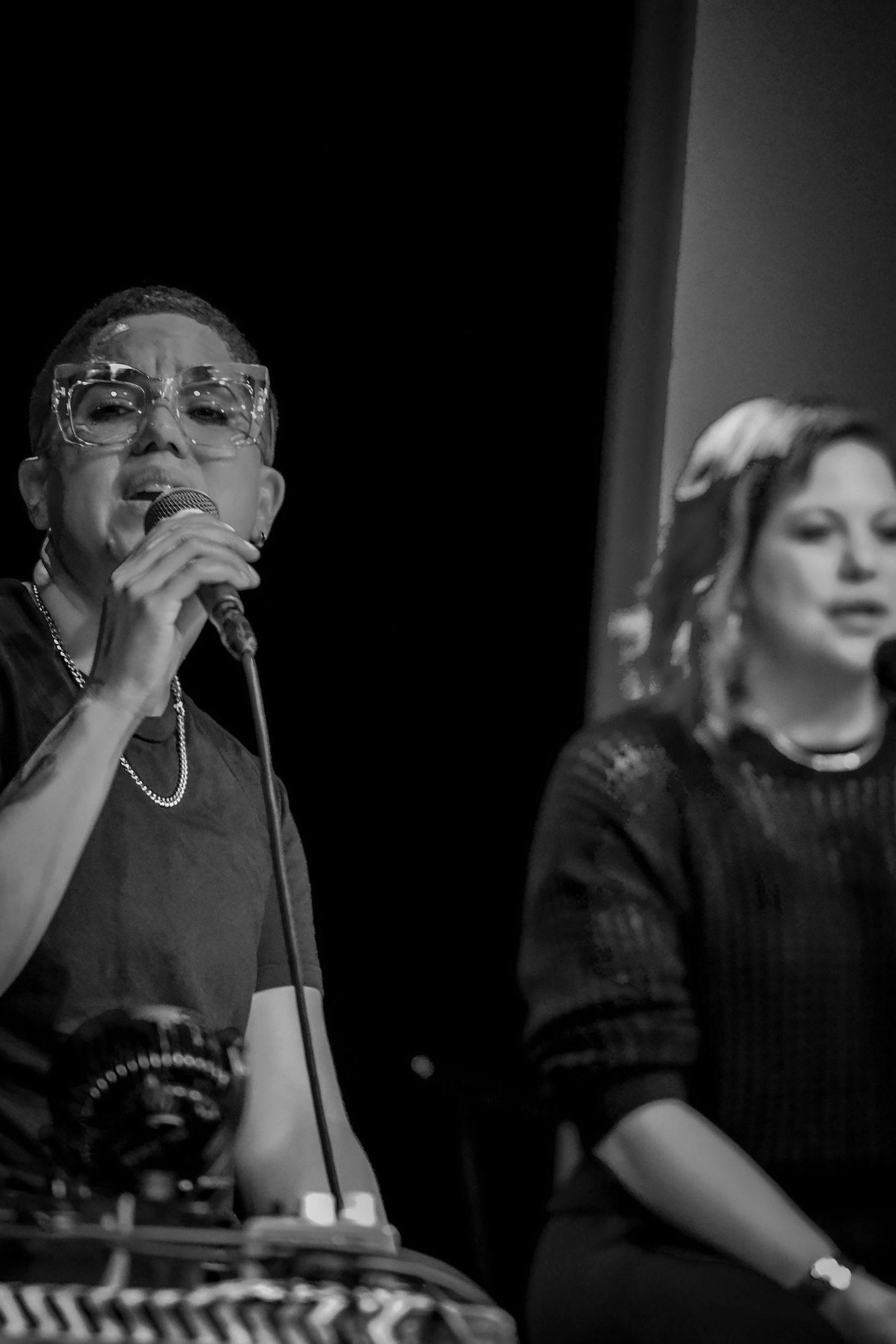 Male vocalist with glasses singing into microphone with female performer in background.