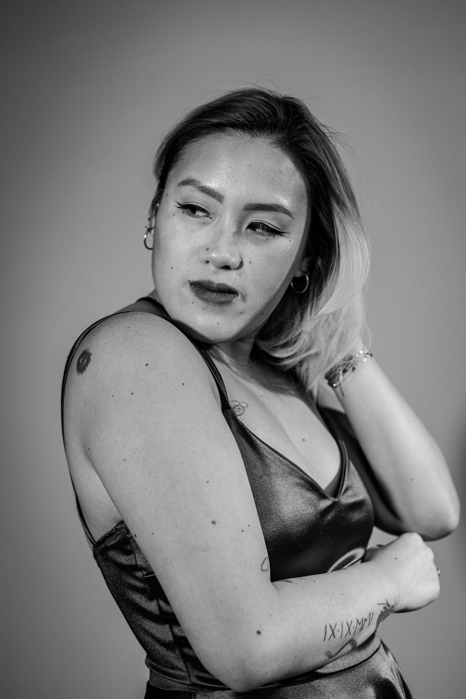 Black and white portrait of a young woman with blonde highlights, tattoos, and bold makeup posing confidently.
