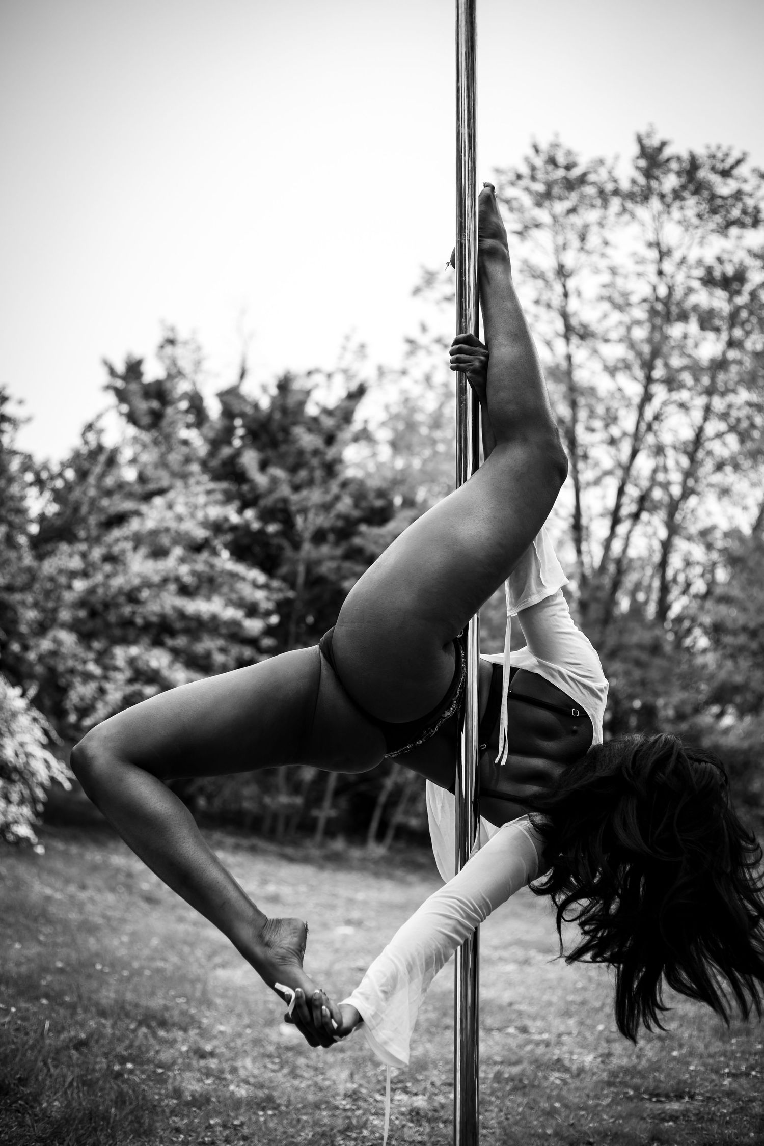 Black and white photo of athletic woman performing inverted pole dance move outdoors.