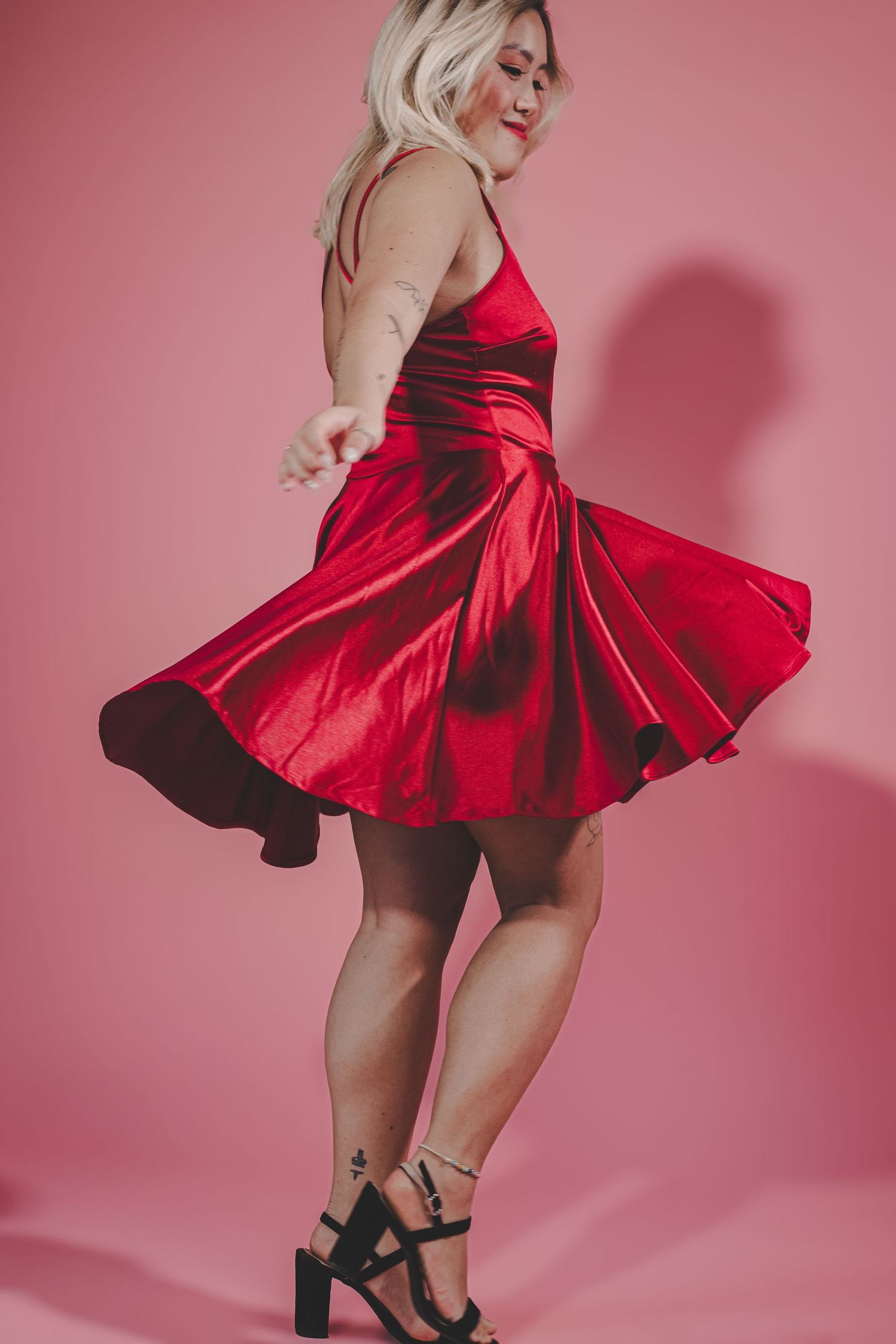 Blonde woman twirling in a shiny red satin mini dress with black heels on pink background.
