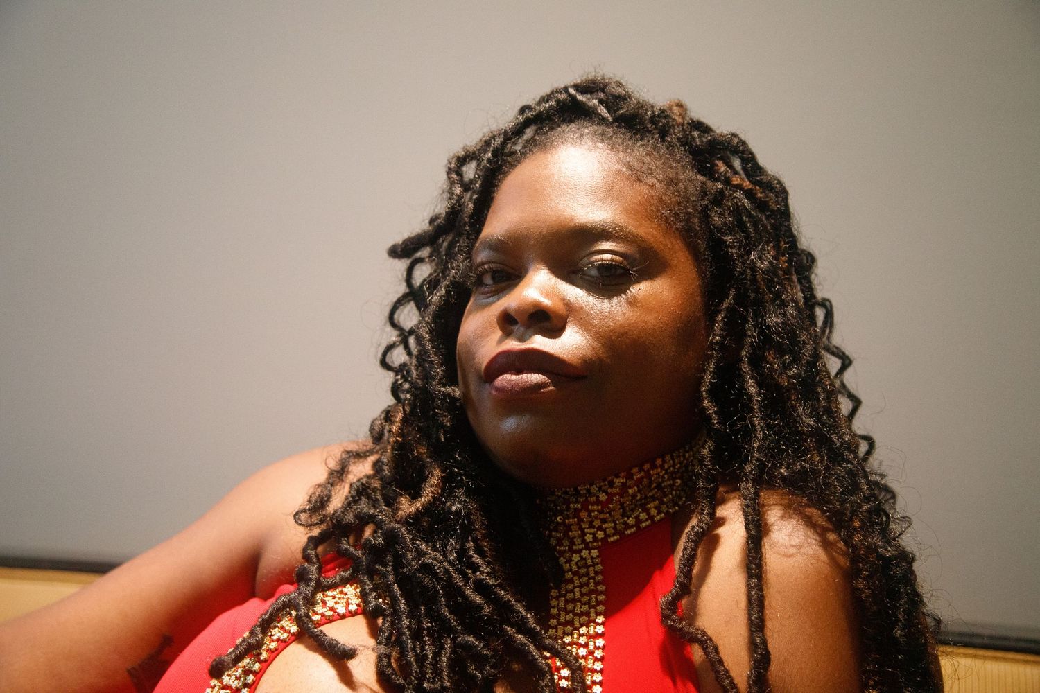 Close-up portrait of a Black woman with long curly locs and layered beaded necklaces in red top.
