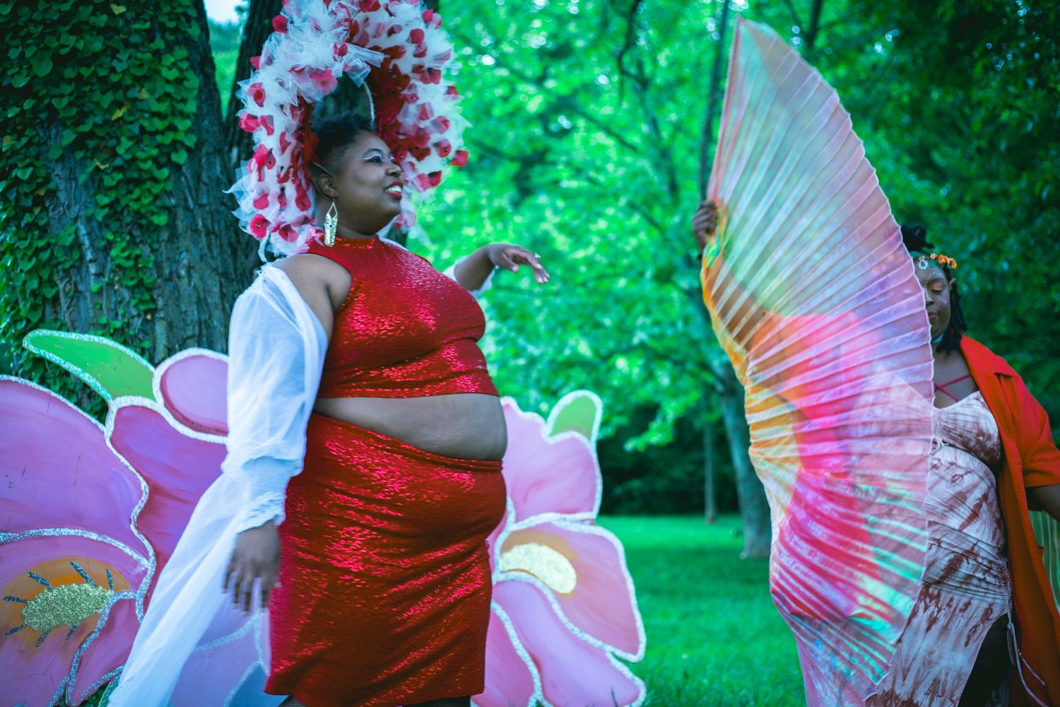 Woman in red sequined dress poses outdoors near a tree with large colorful butterfly wings and pink floral backdrop.