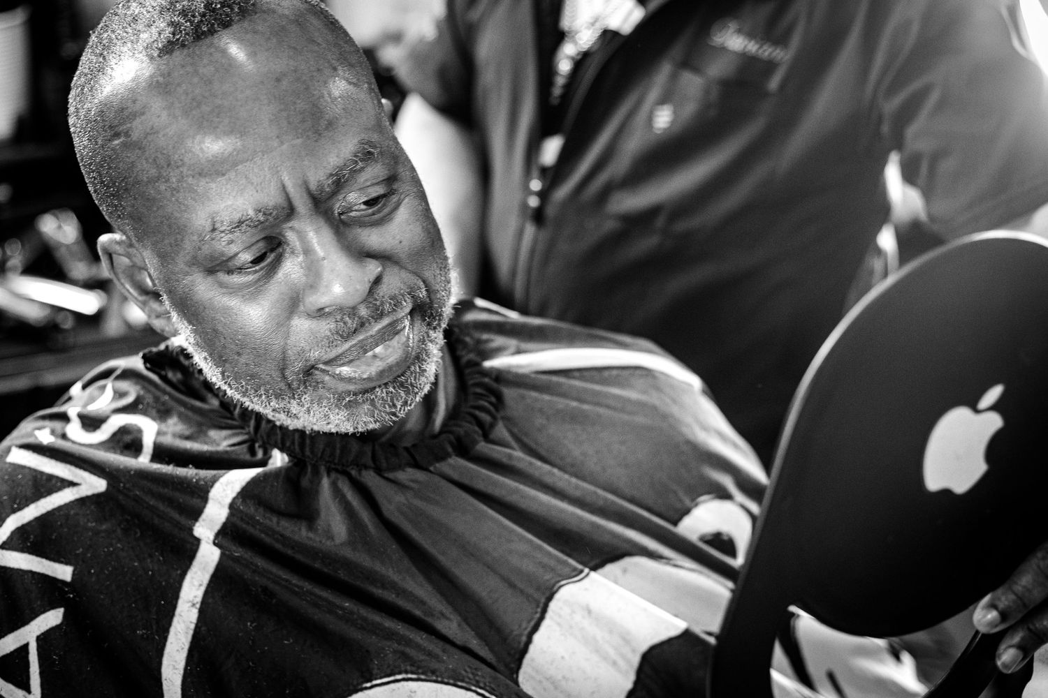 Black and white close-up of a man examining his haircut using an Apple-branded hand mirror.