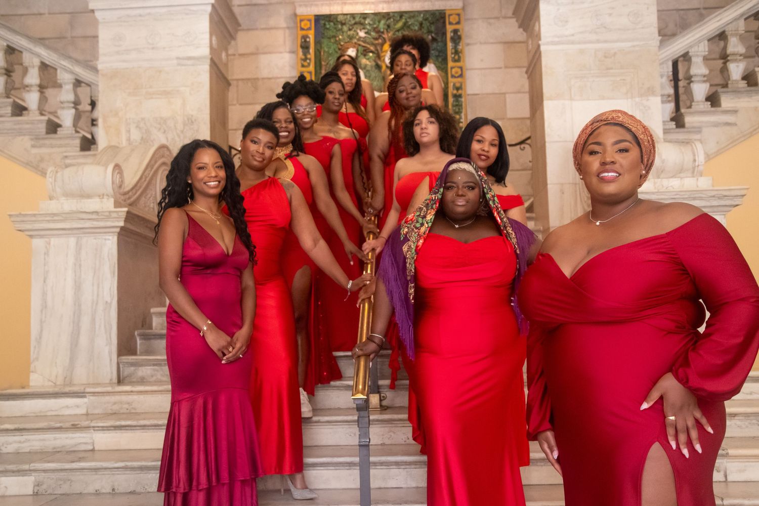 Black Women in red and pink formal gowns posing together on ornate marble staircase.