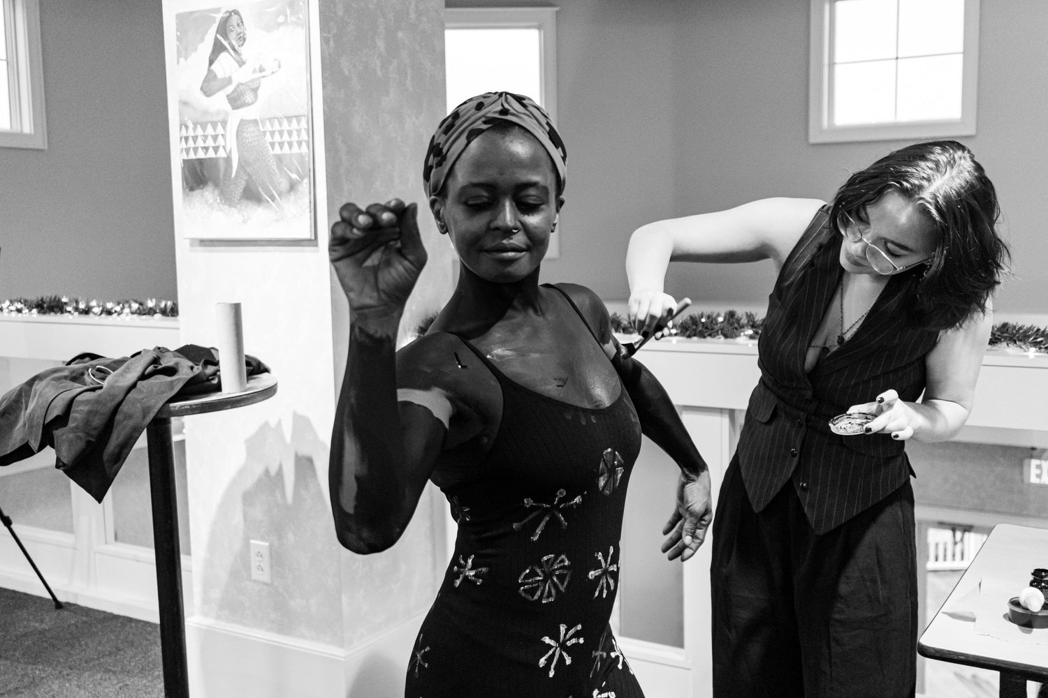 Black and white photo of a woman in a floral dress posing while another adjusts her outfit backstage.
