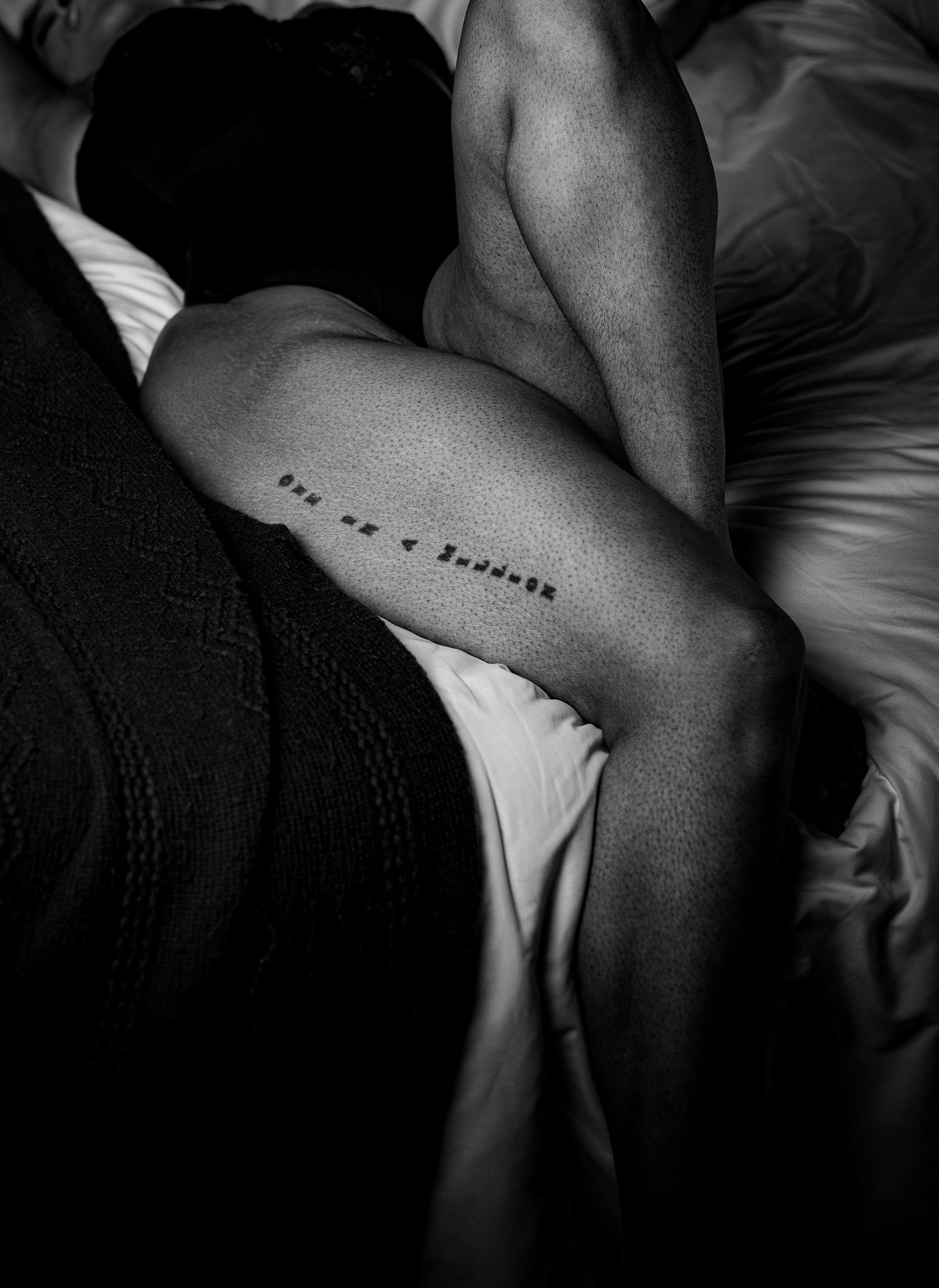 Black and white close-up of a leg tattoo reading on a bed with white sheets.