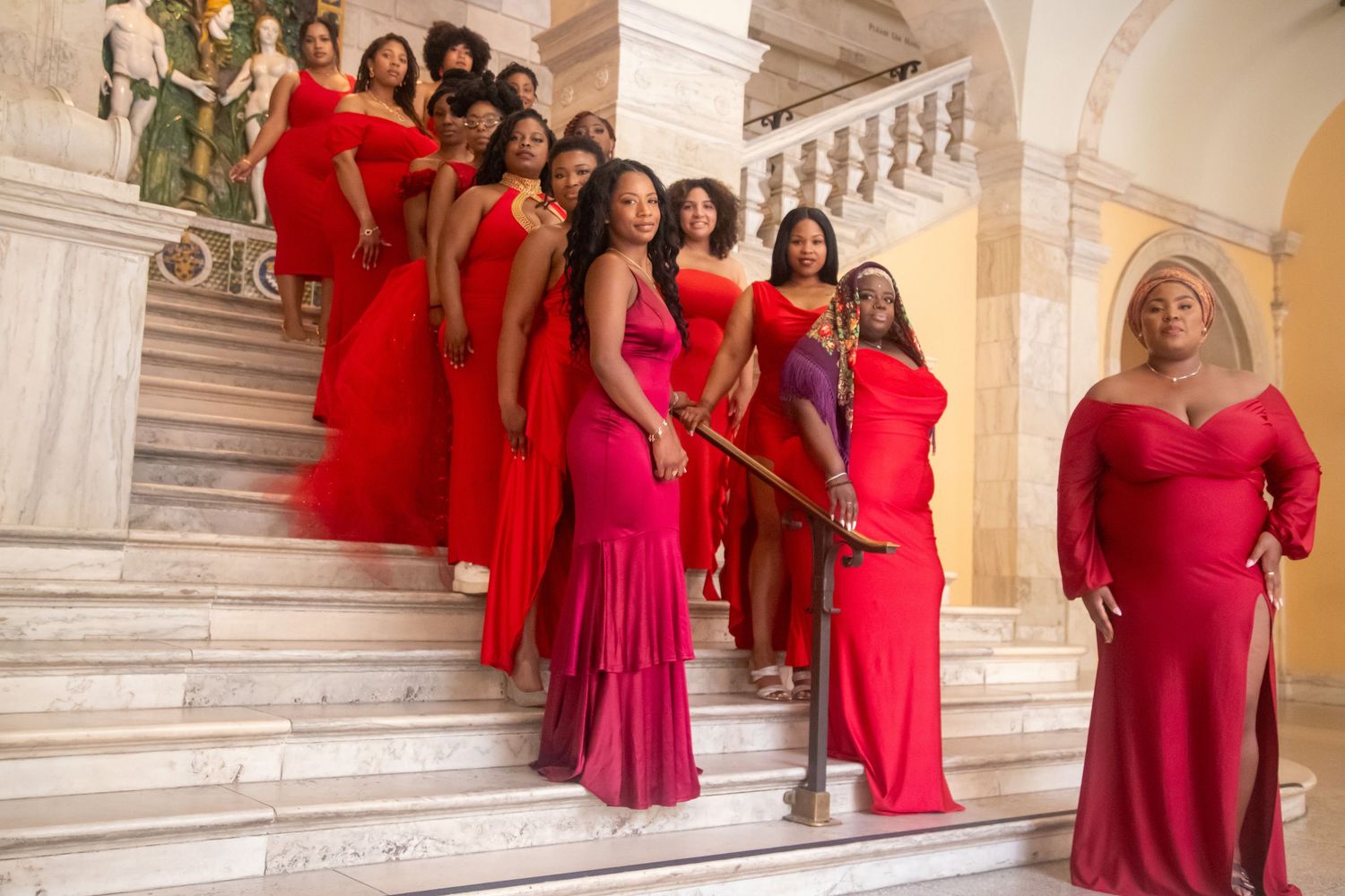 Black Women in vibrant red and pink formal dresses posing on luxurious marble staircase.