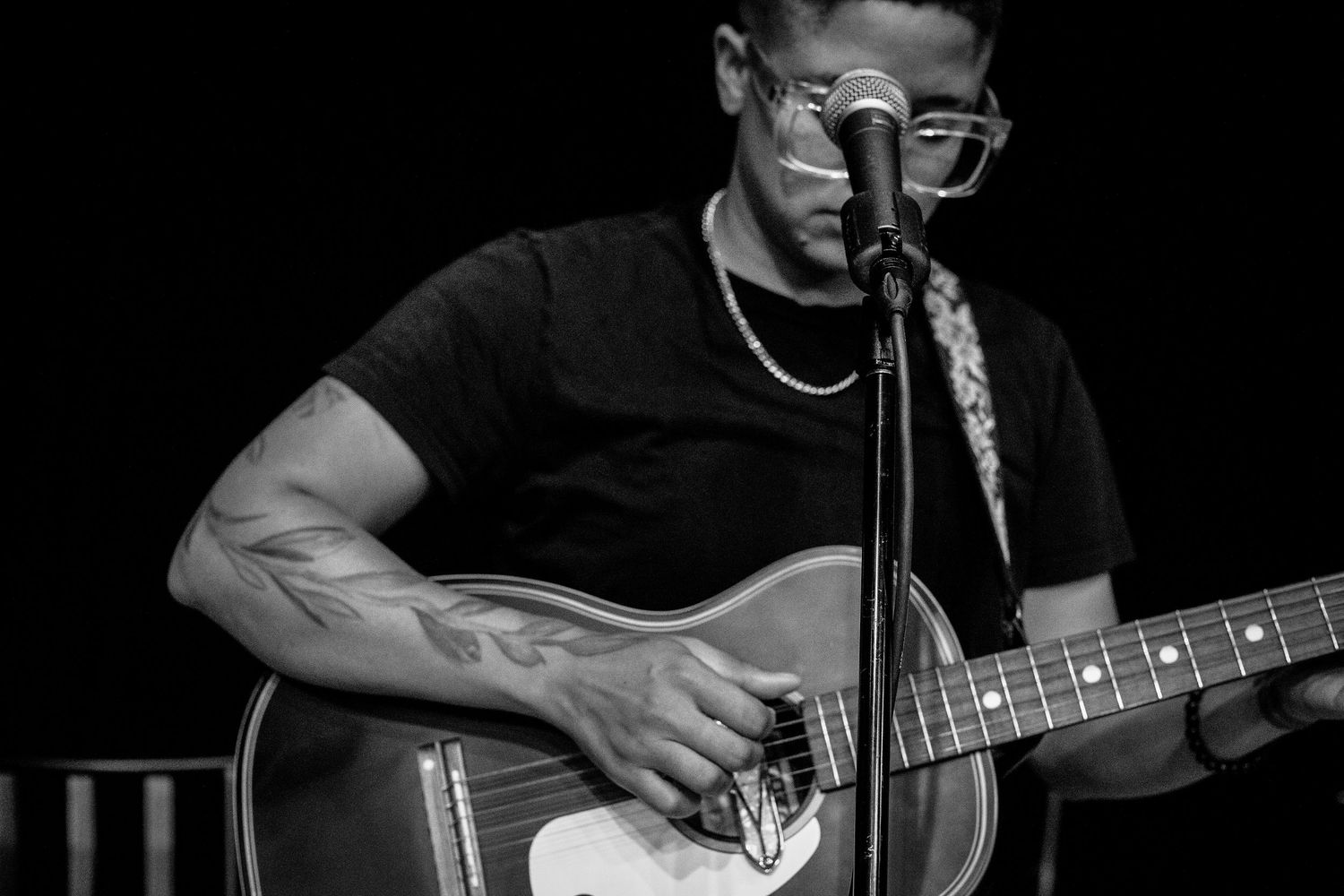 Black and white photo of a tattooed musician playing acoustic guitar and singing into a microphone on stage.