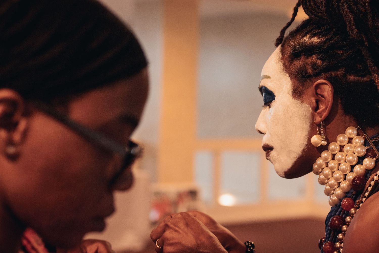 Makeup artist applies detailed white face paint to a performer adorned with pearl necklaces in profile view.