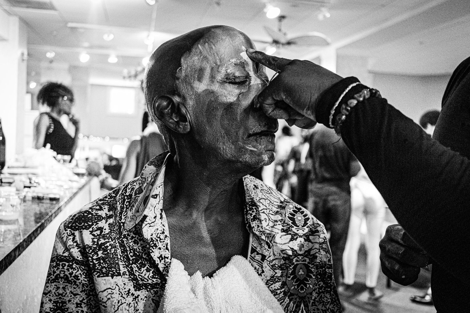 Black and white photo of makeup artist applying face paint to a model backstage.