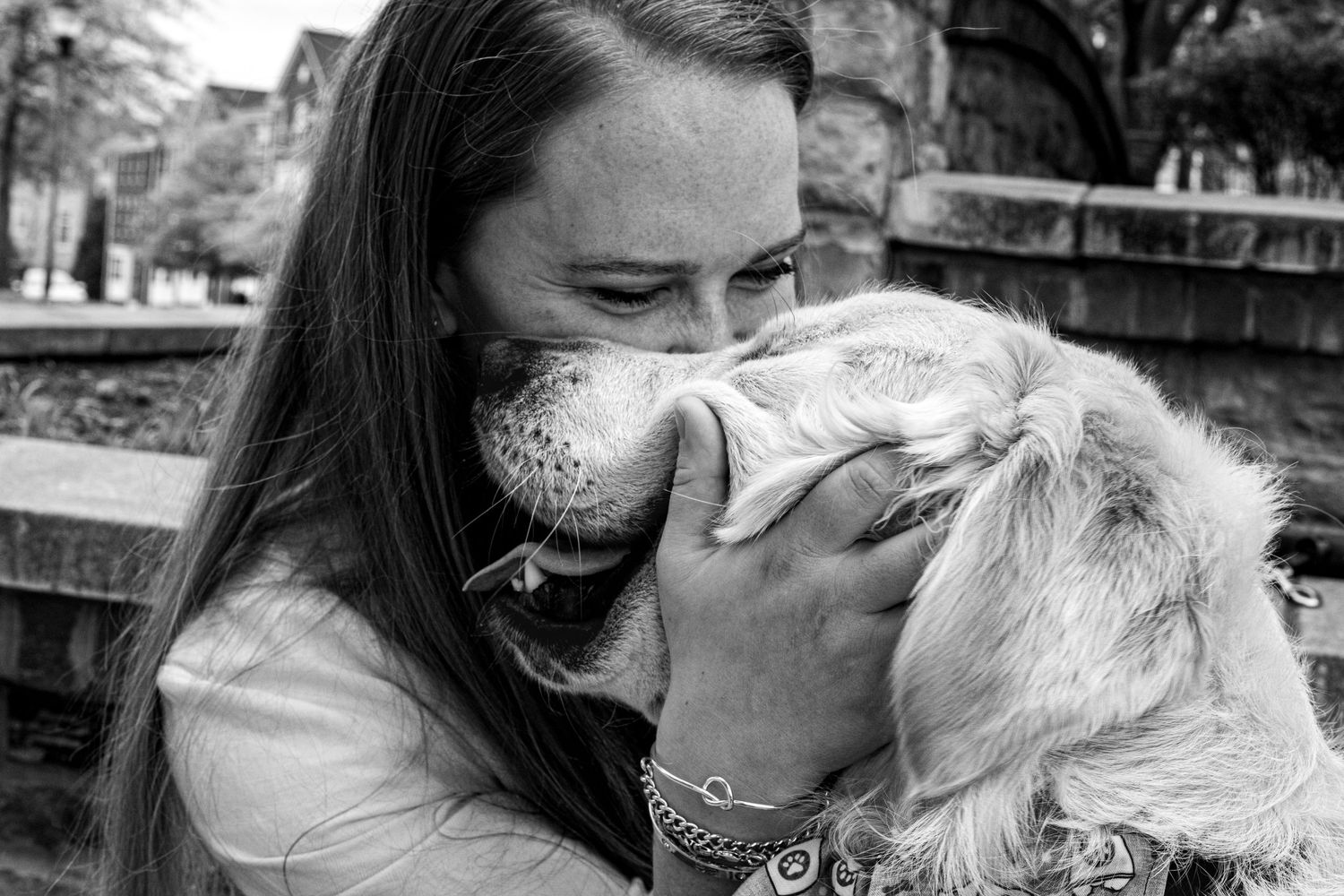 Black and white photo of a woman outdoors smiling and lovingly holding and kissing a fluffy golden retriever's face as it nuzzles her face.