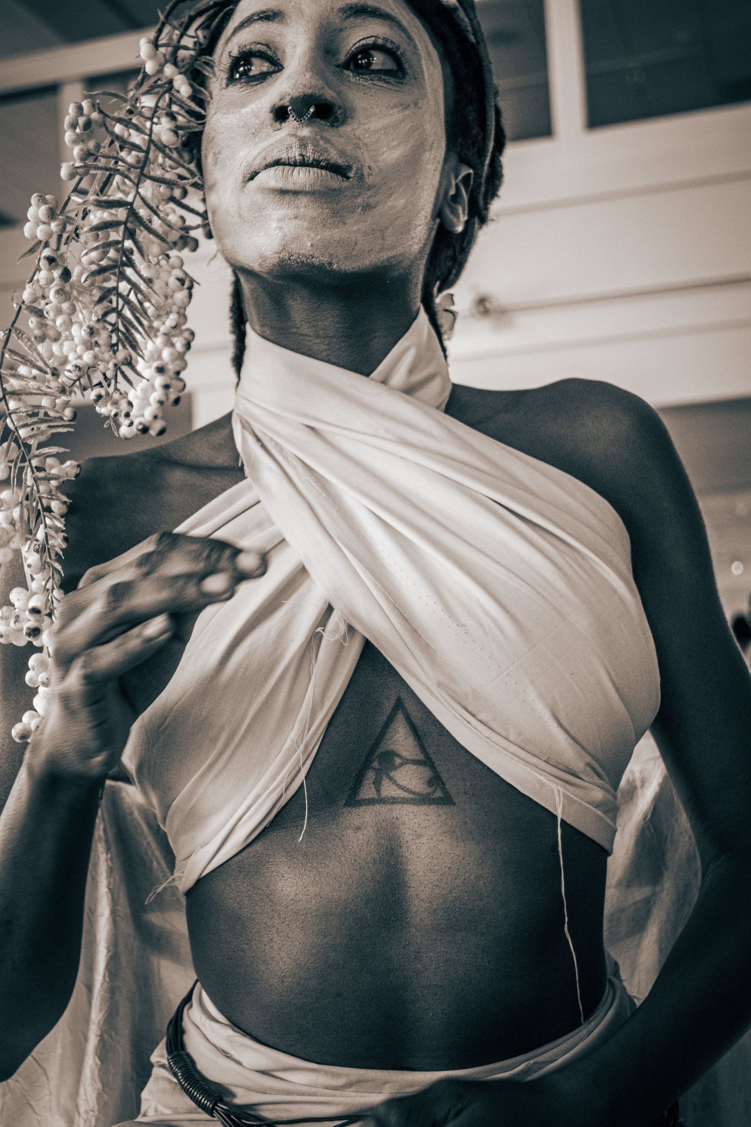 Sepia toned portrait of a model with white face paint in draped white fabric with Eye of Horus chest tattoo.