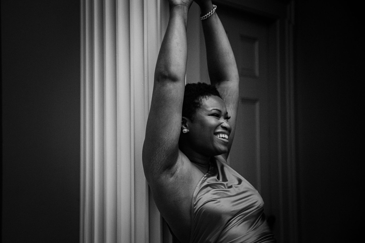 Black and white photo of a woman stretching arms overhead beside a classical column, smiling.