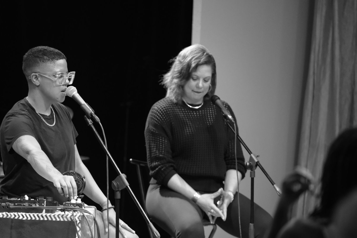 Black and white image of two women seated on stage, both speaking into microphones at a live event.