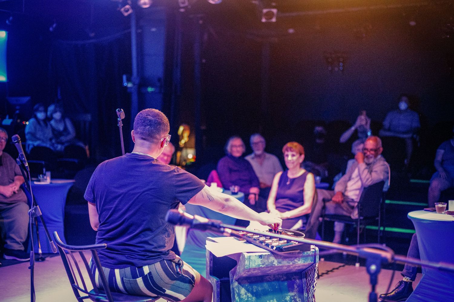 Musician performs on keyboard for small intimate audience in colorful blue-lit venue setting.