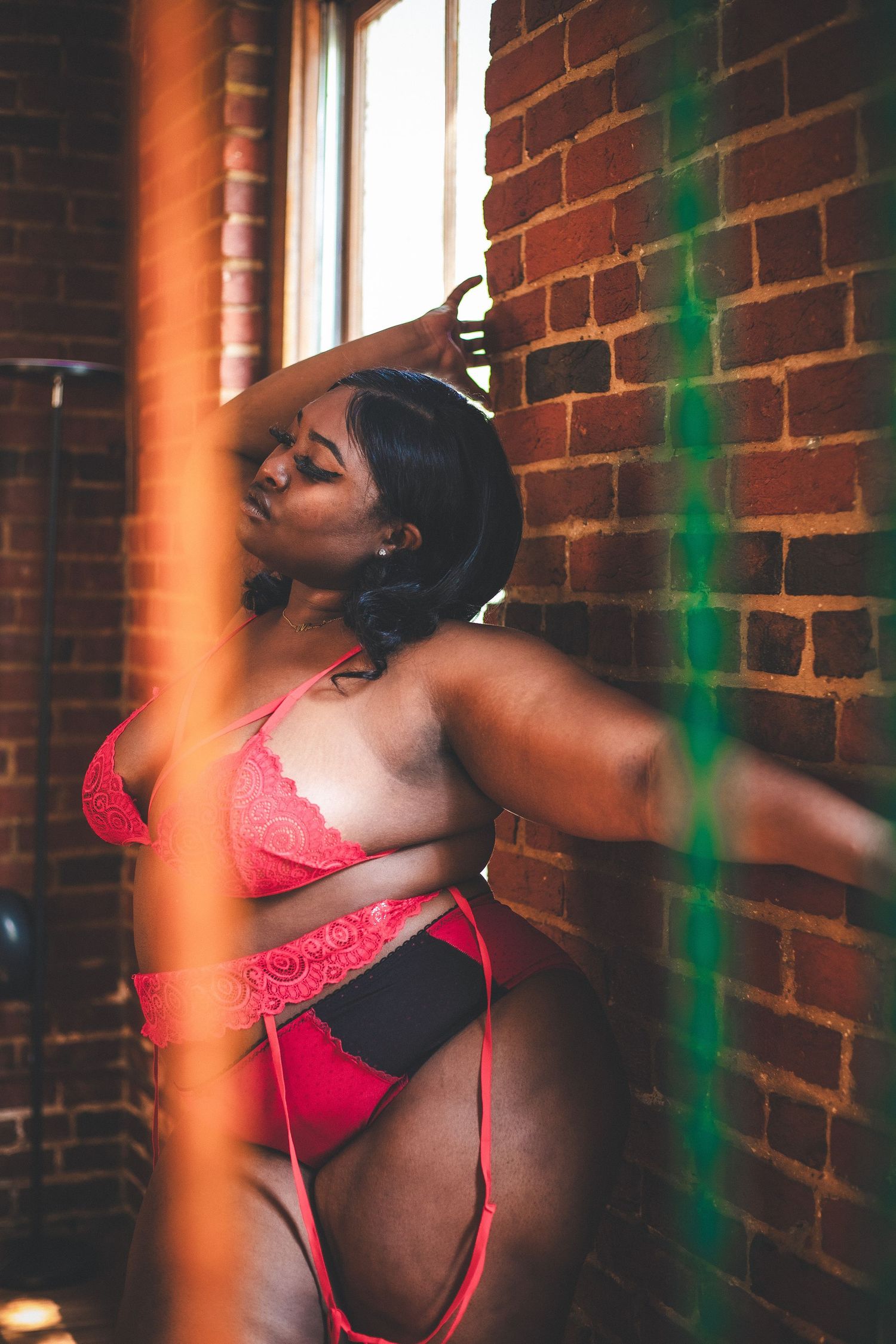 Curvy woman in red lace lingerie posing confidently by a window against an exposed brick wall.